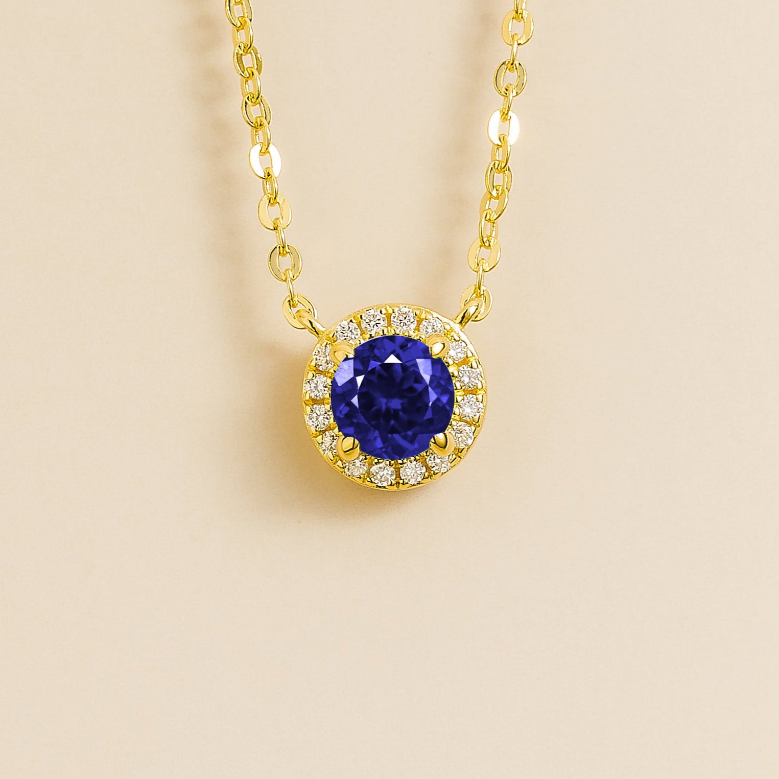 A gold necklace with a halo design featuring a round blue sapphire centerpiece surrounded by smaller diamonds, displayed against a light background.