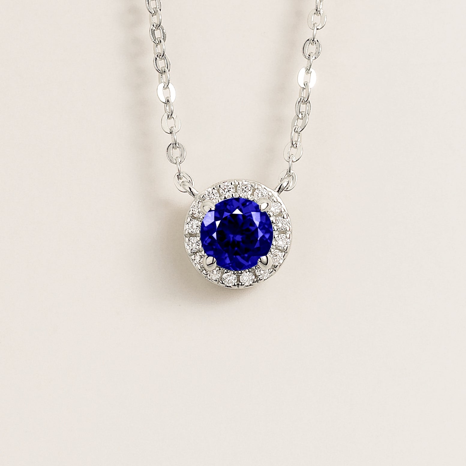 A white gold gold necklace with a halo design featuring a round blue sapphire centerpiece surrounded by smaller diamonds, displayed against a light background.
