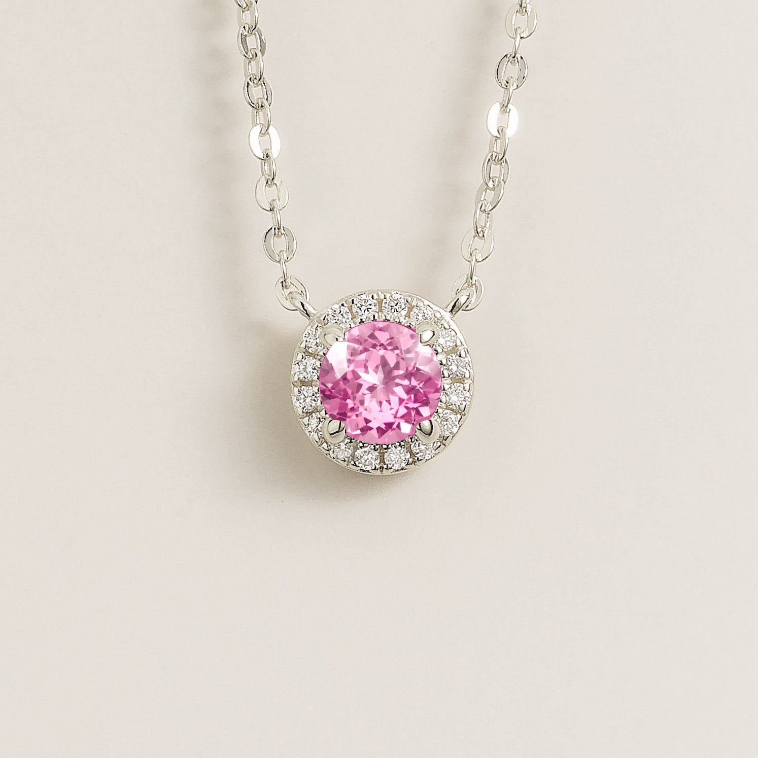 A white gold gold necklace with a halo design featuring a round pink sapphire centerpiece surrounded by smaller diamonds, displayed against a light background.