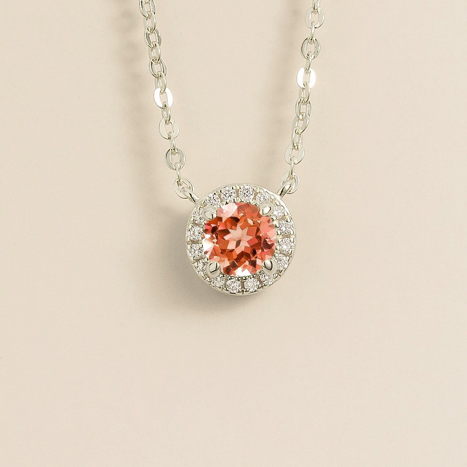 A white gold gold necklace with a halo design featuring a round padparadscha sapphire centerpiece surrounded by smaller diamonds, displayed against a light background.