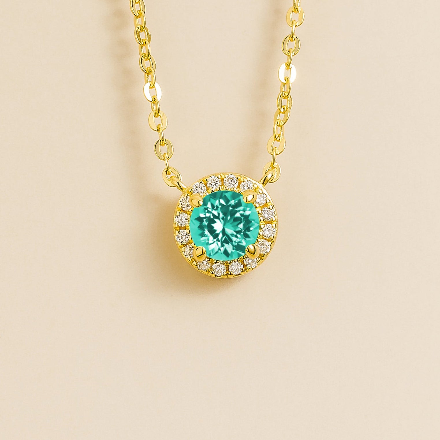A gold necklace with a halo design featuring a round paraiba sapphire centerpiece surrounded by smaller diamonds, displayed against a light background.