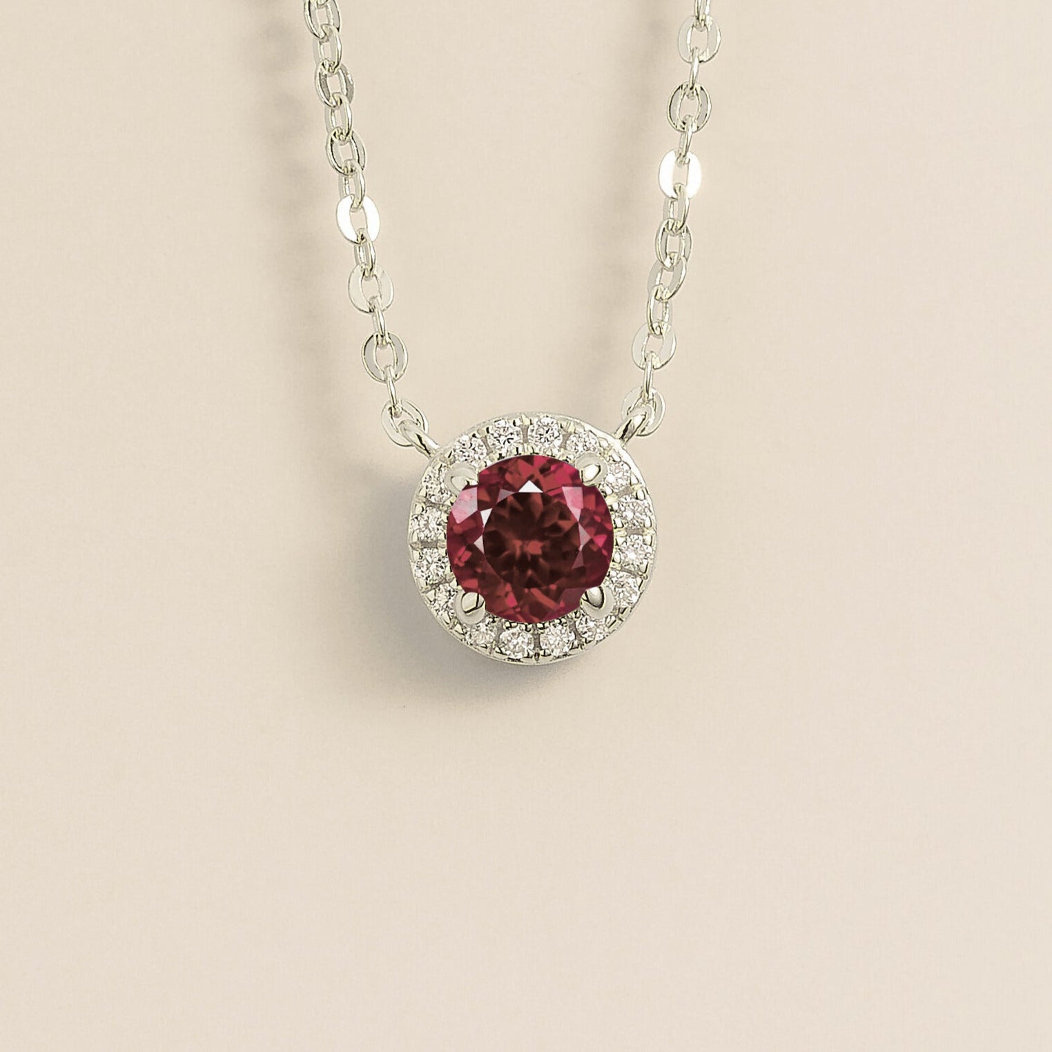 A white gold gold necklace with a halo design featuring a round ruby centerpiece surrounded by smaller diamonds, displayed against a light background.