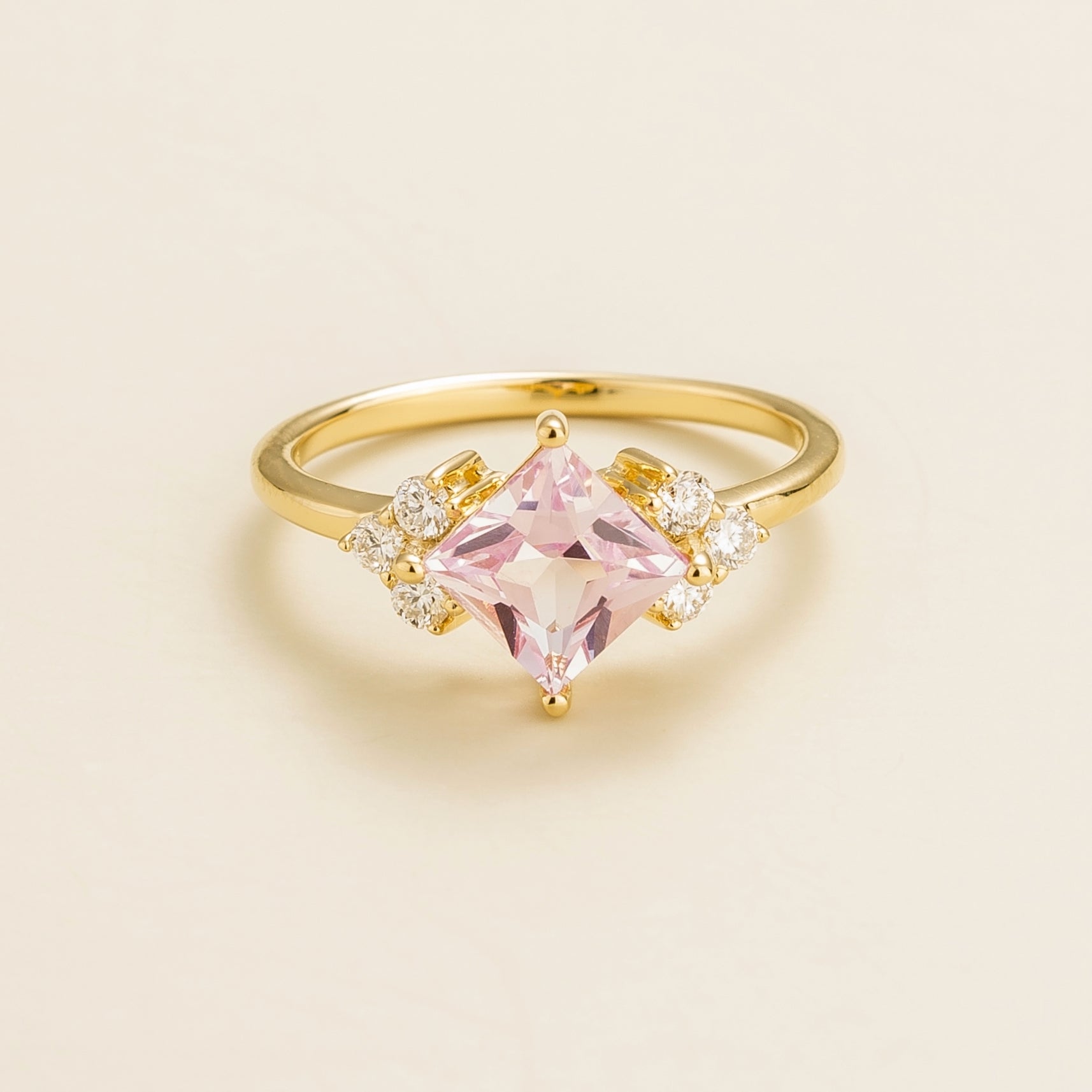 A gold ring with a princess cut pink sapphire center stone and surrounding diamonds, set on a plain background.
