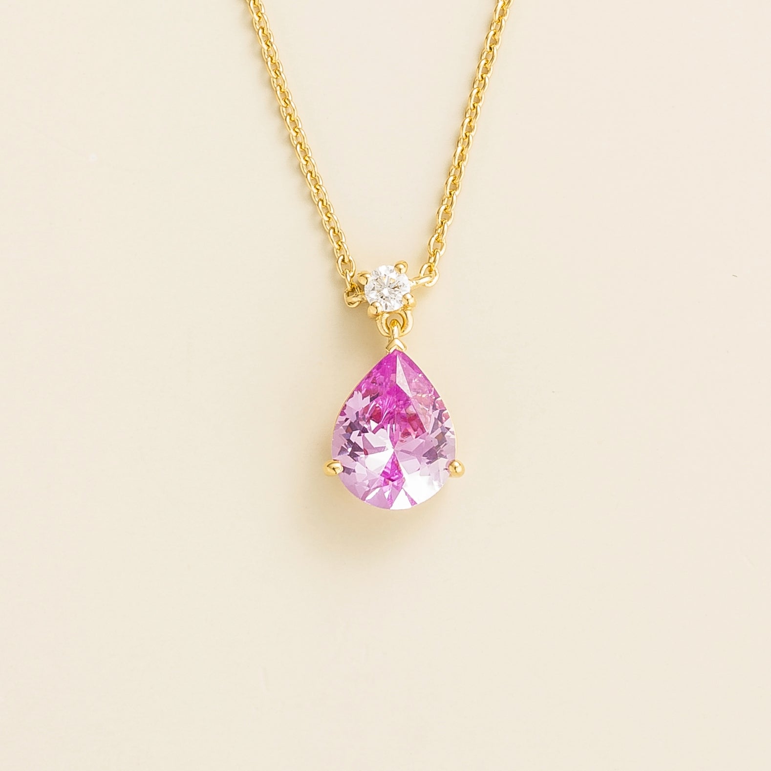A classic gold necklace with a pear shape pink sapphire pendant and a smaller round diamond, displayed against a neutral background.