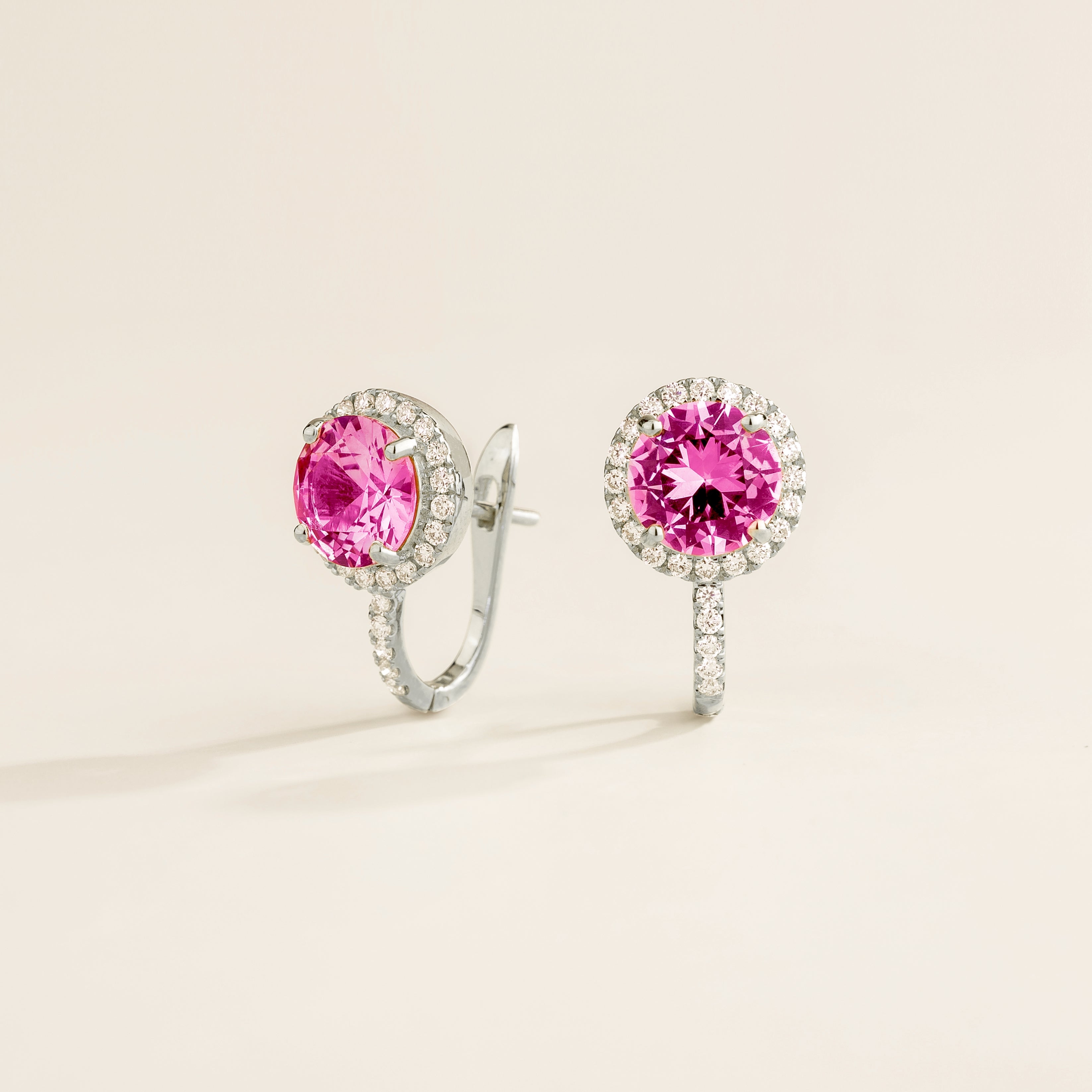 Pair of white gold earrings with round pink sapphire gemstones and diamond halo on a light background