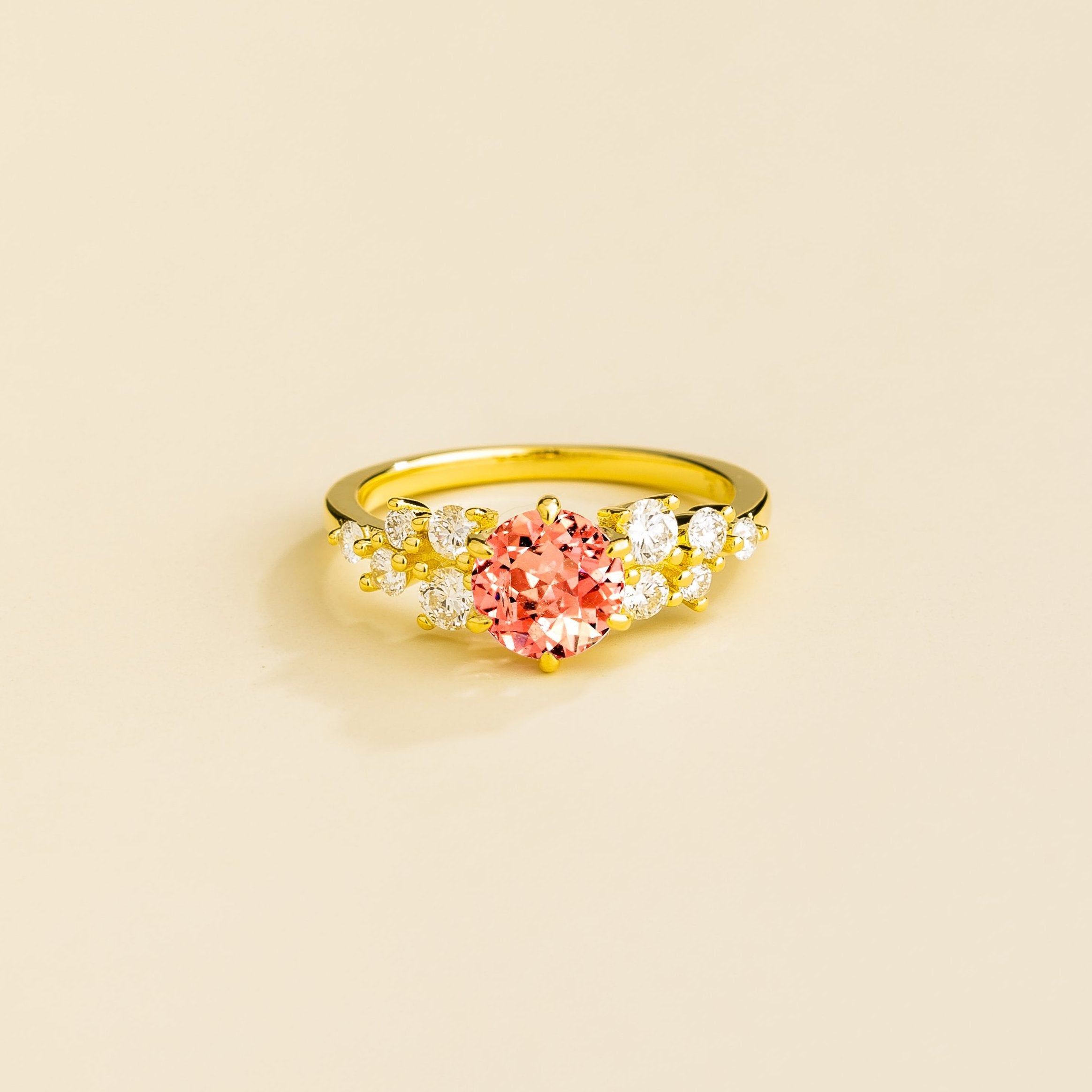 A gold ring with a padparadscha sapphire center stone and smaller diamonds cluster set in gold, displayed against a neutral background.