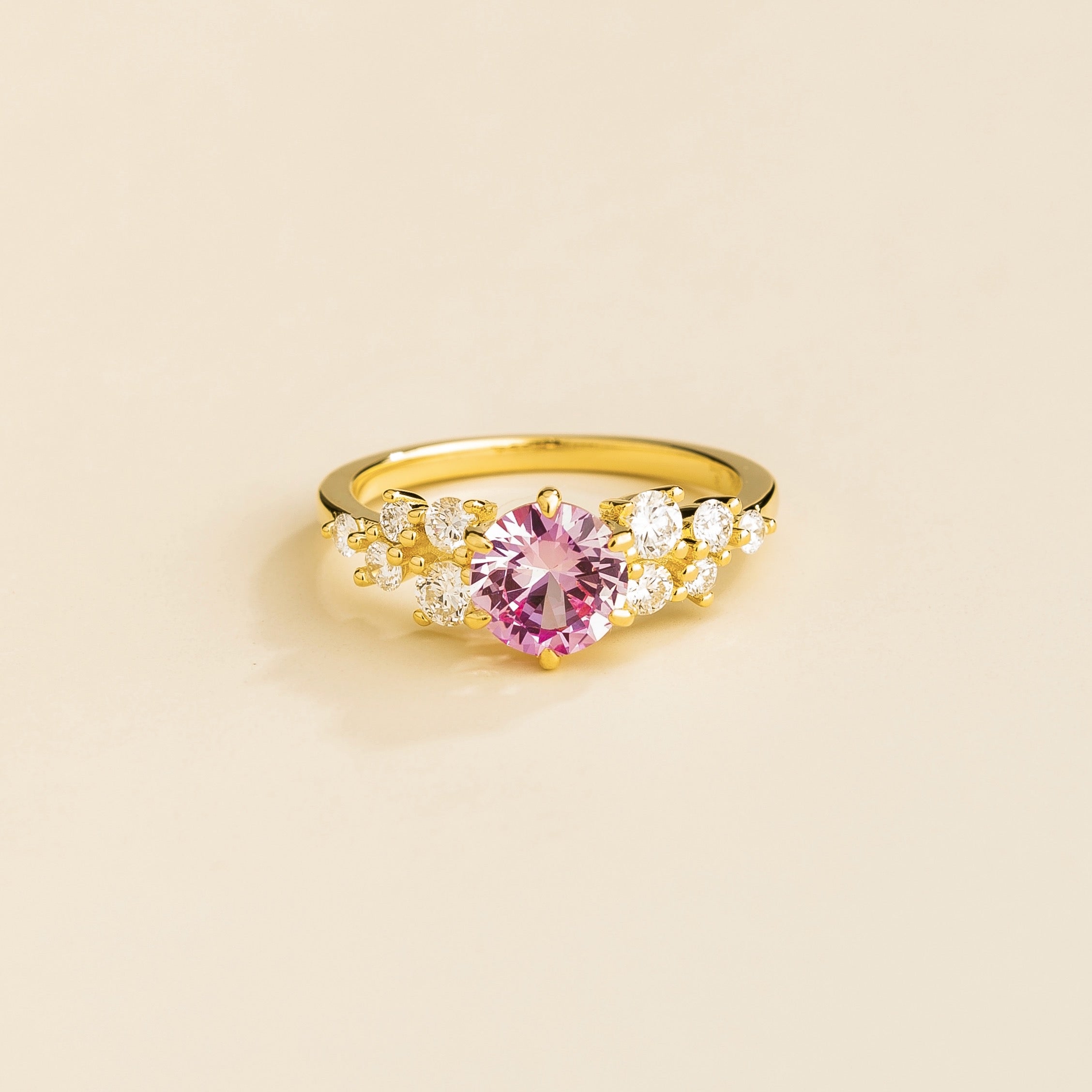 A gold ring with a pink sapphire center stone and smaller diamonds cluster set in gold, displayed against a neutral background.