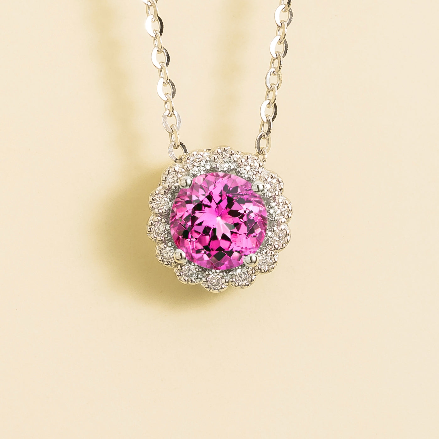 A white gold necklace set with a round pink sapphire pendant surrounded by smaller halo diamonds, featuring an adjustable chain.