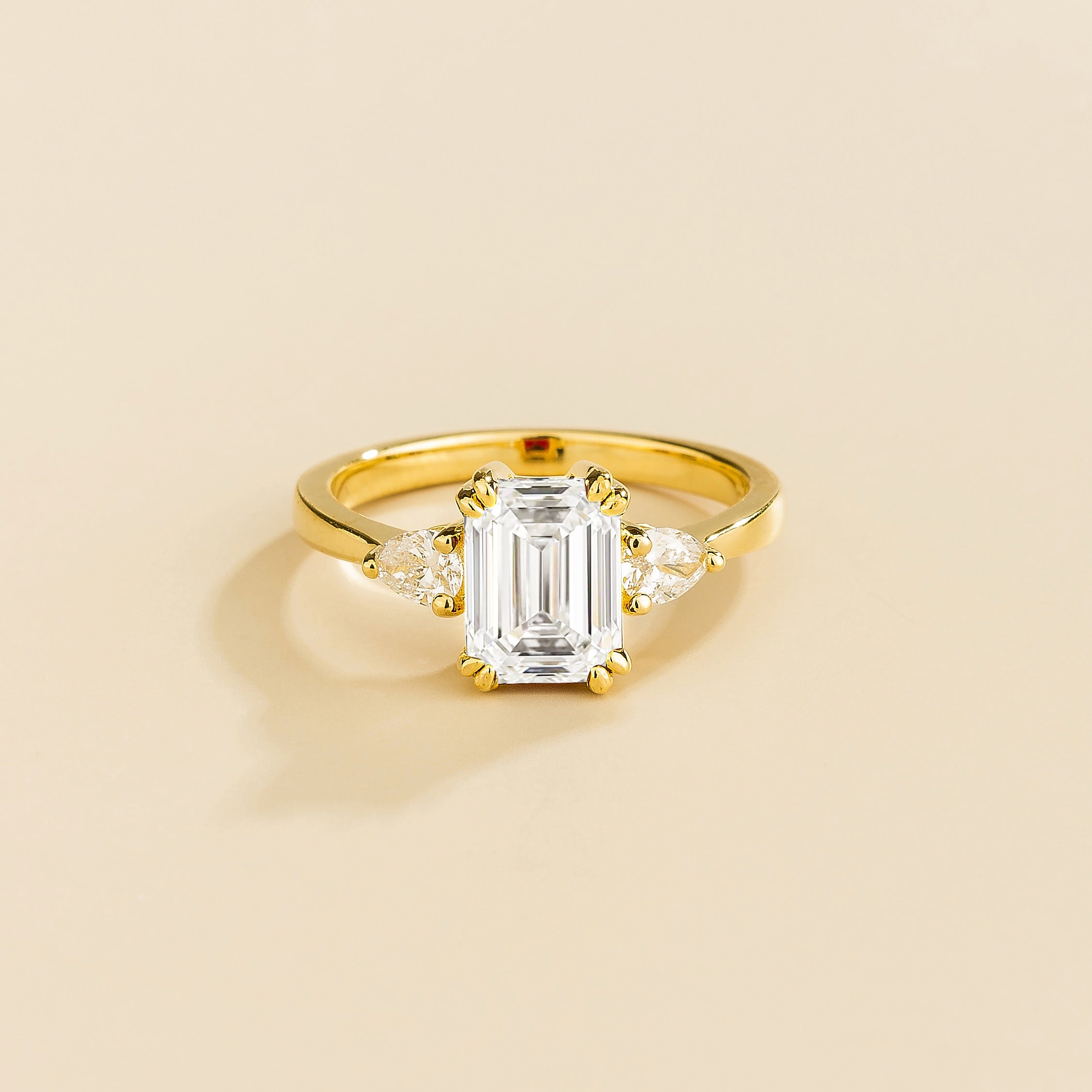 Diamond ring with emerald-cut center stone and pear side diamonds set in gold on a light beige background
