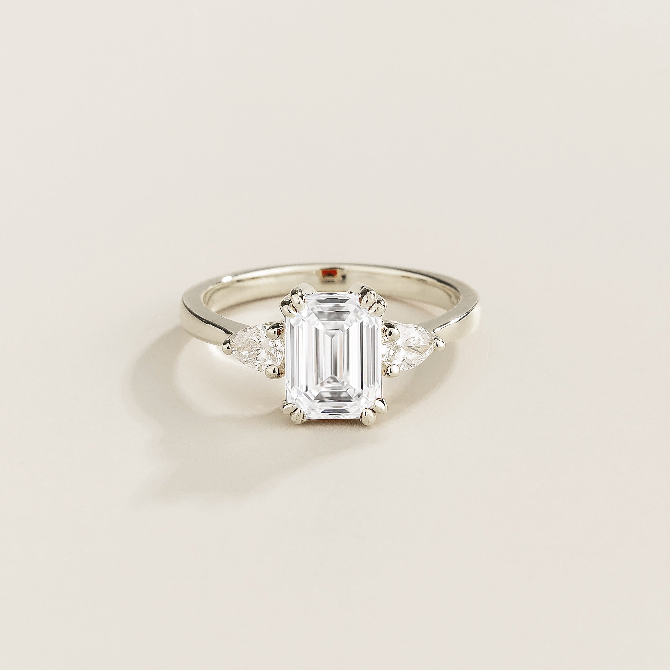 Diamond ring with emerald-cut center stone and pear side diamonds set in white gold on a light beige background