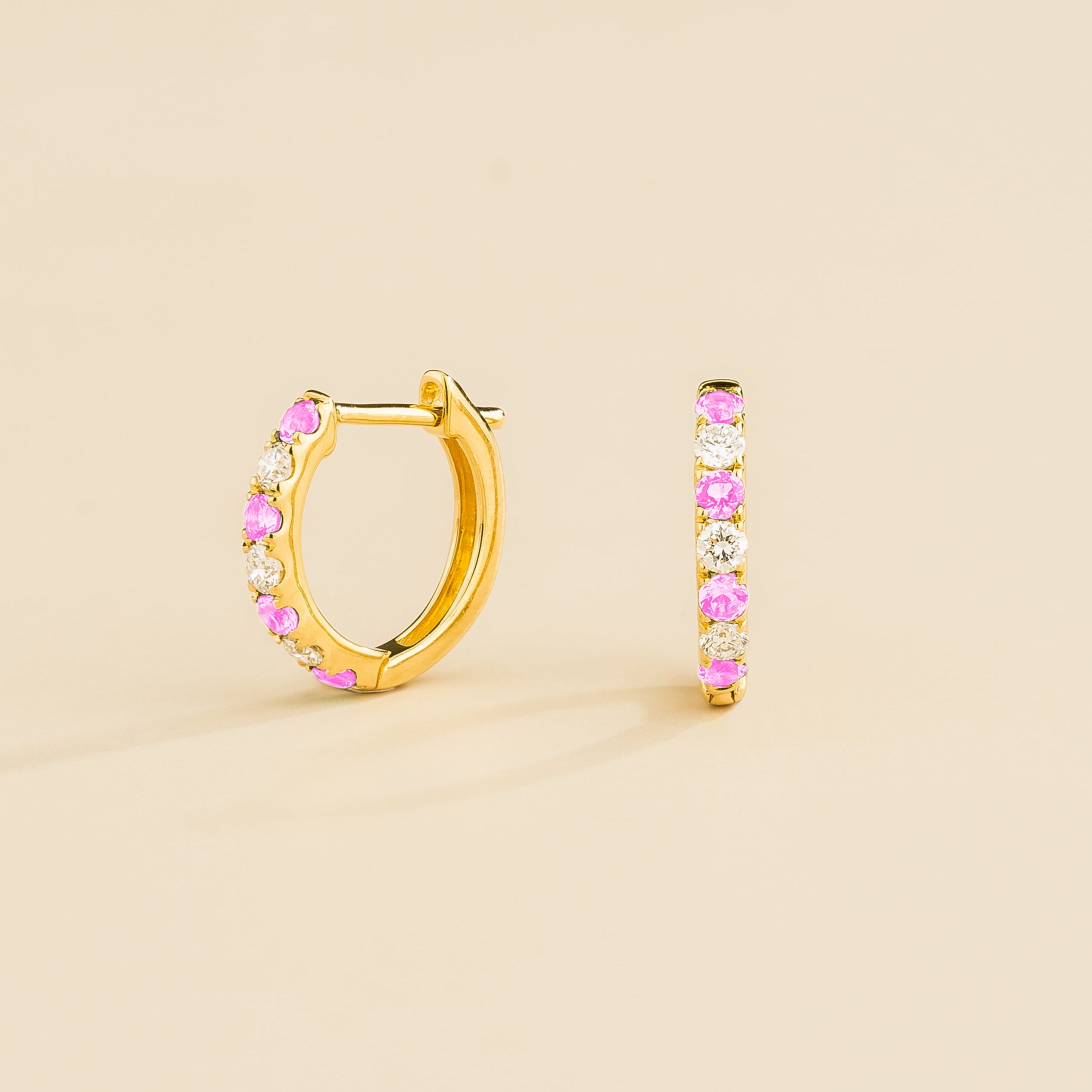 A pair of gold huggie earrings set with pink sapphire and diamonds on a beige background.