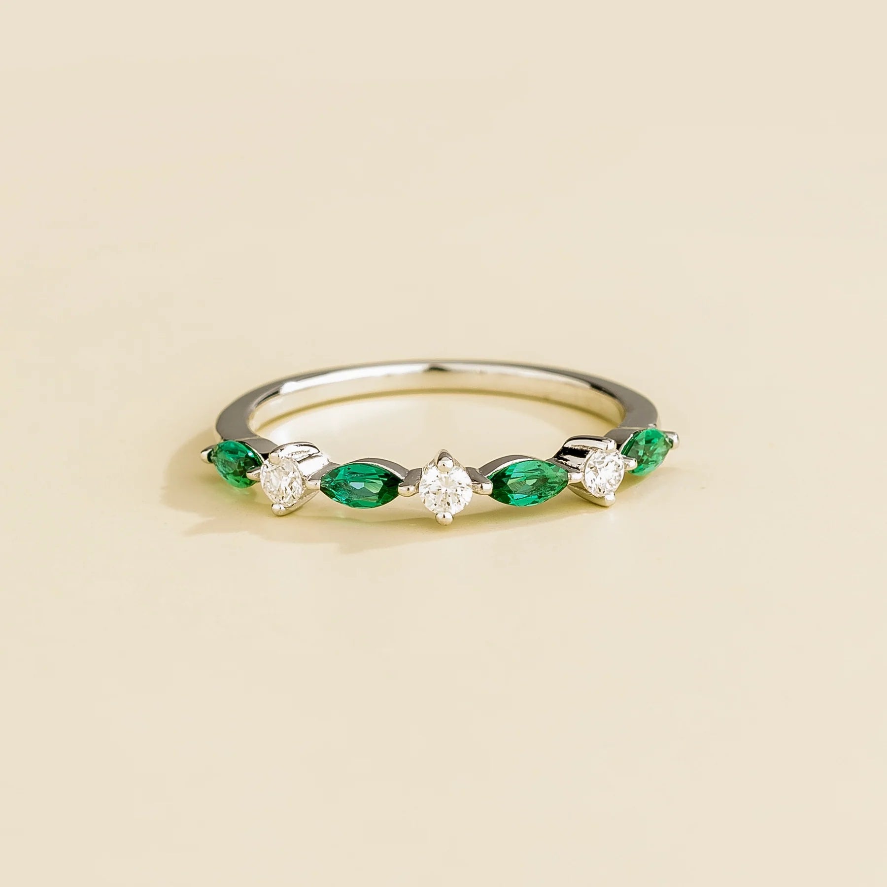 A white gold band ring with marquise cut emerald and round cut diamonds set in an alternating pattern on a plain background.