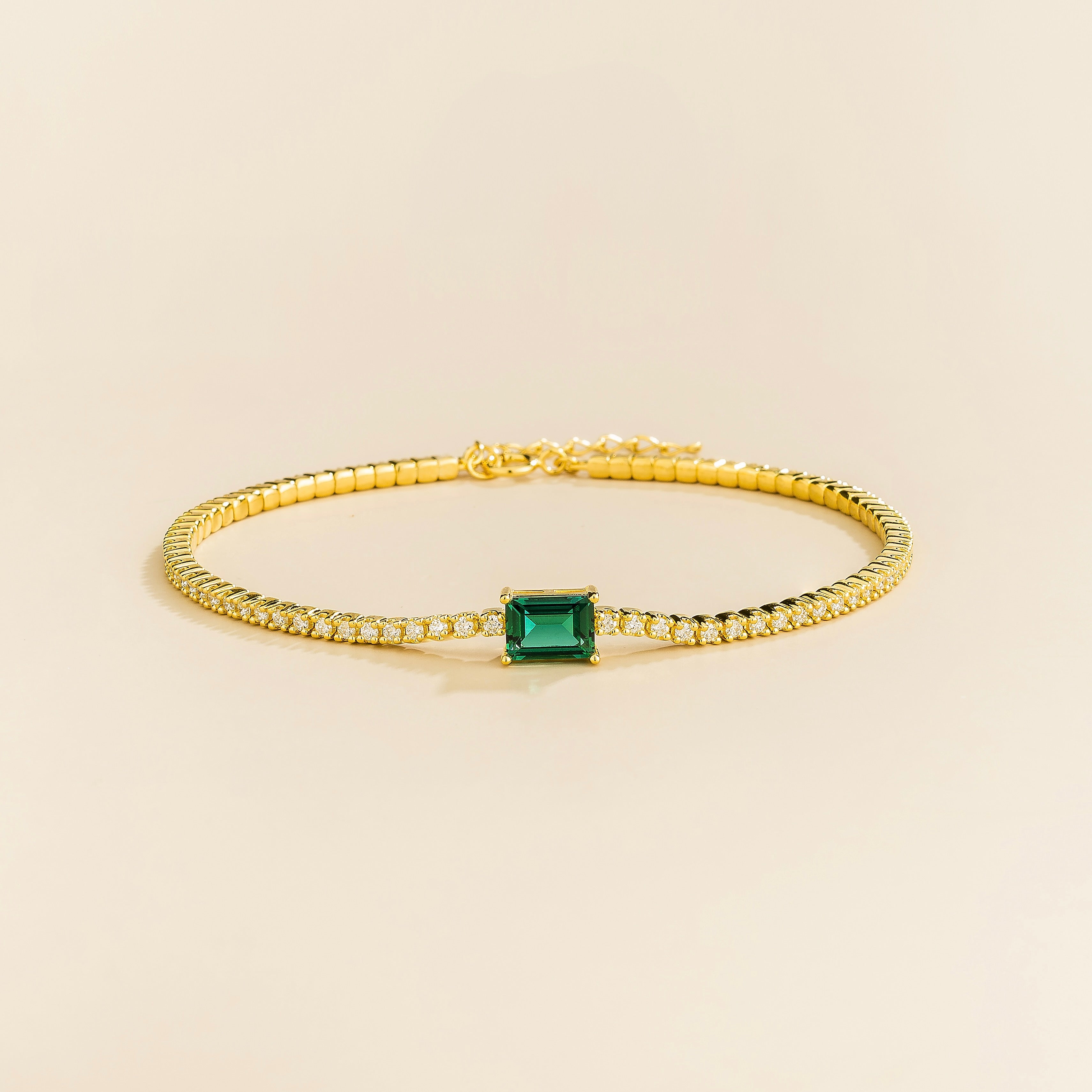 A gold tennis bracelet featuring a central emerald cut diamond with smaller round diamonds around it, displayed against a neutral background.