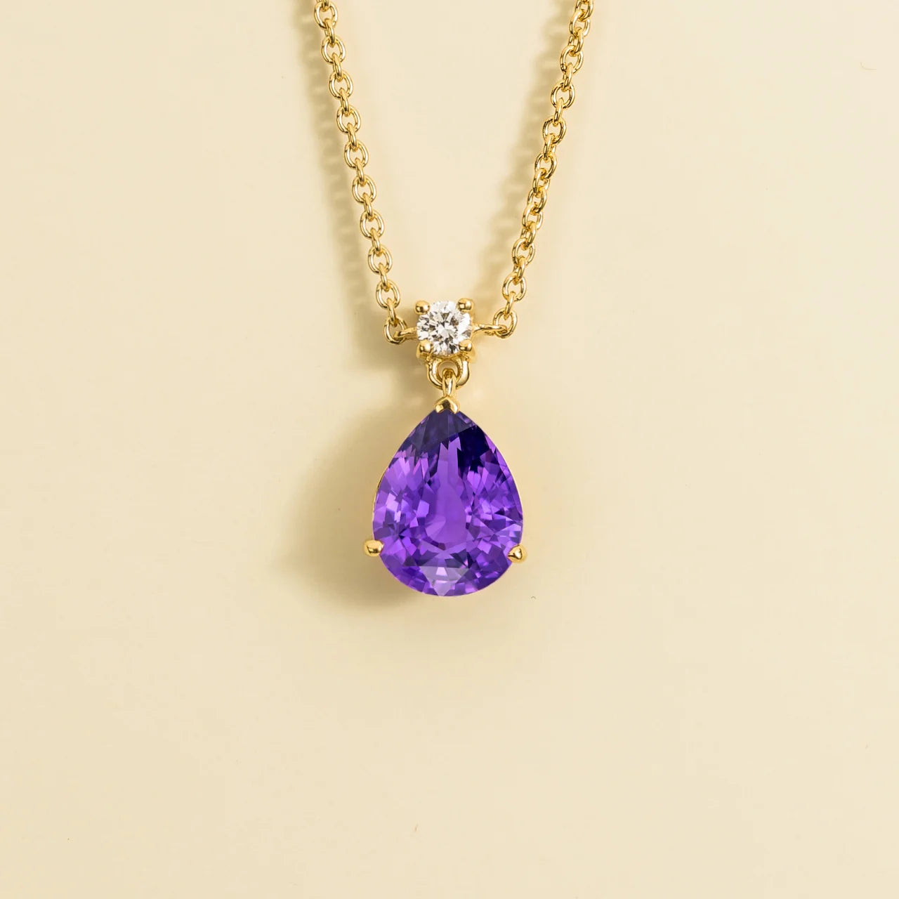 A classic pendant necklace with a pear shape purple sapphire and round diamond set in gold, displayed against a neutral background. By Bespoke Jewellery London