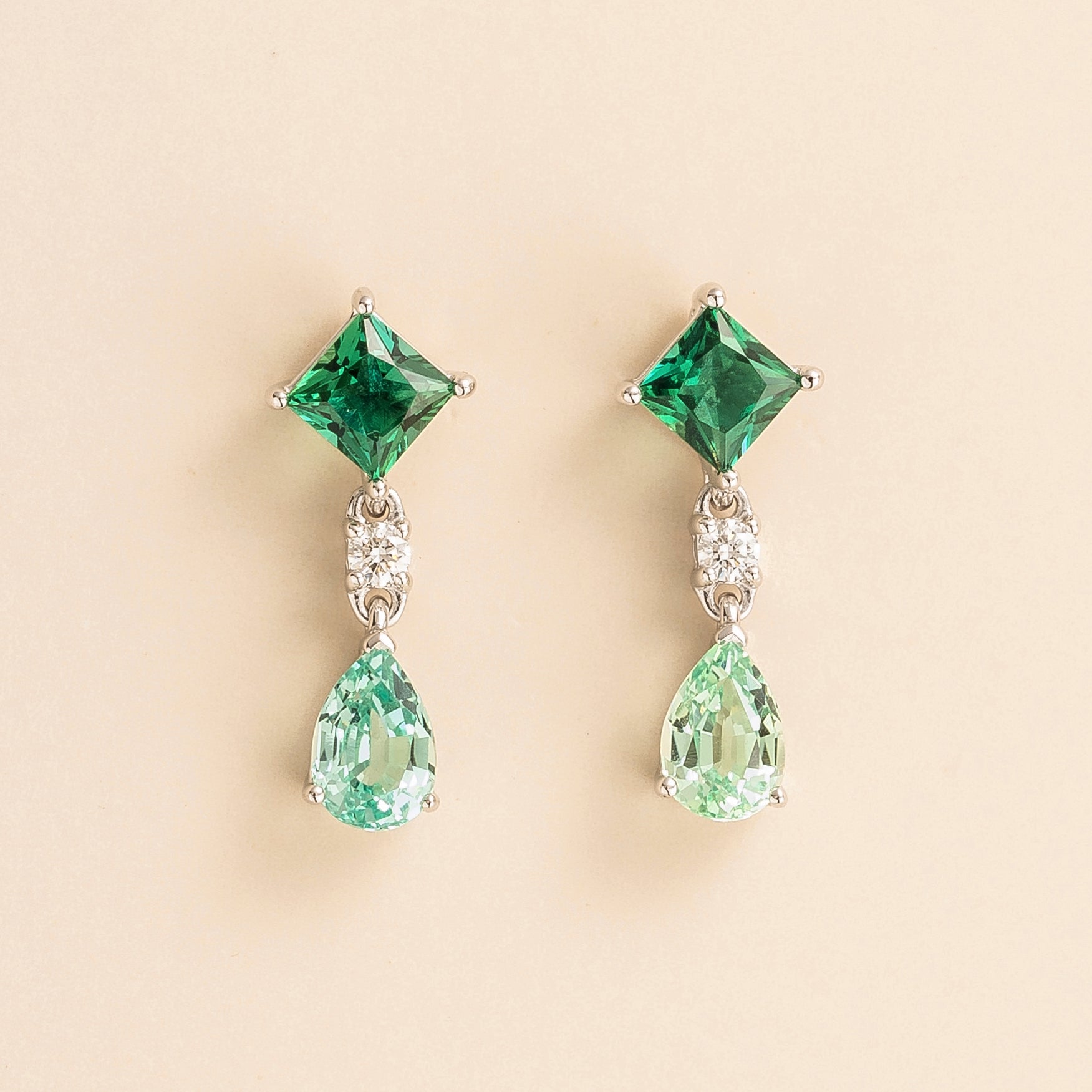 A pair of white gold earrings featuring princess cut emeralds, round cut diamonds, and pear cut green sapphires.