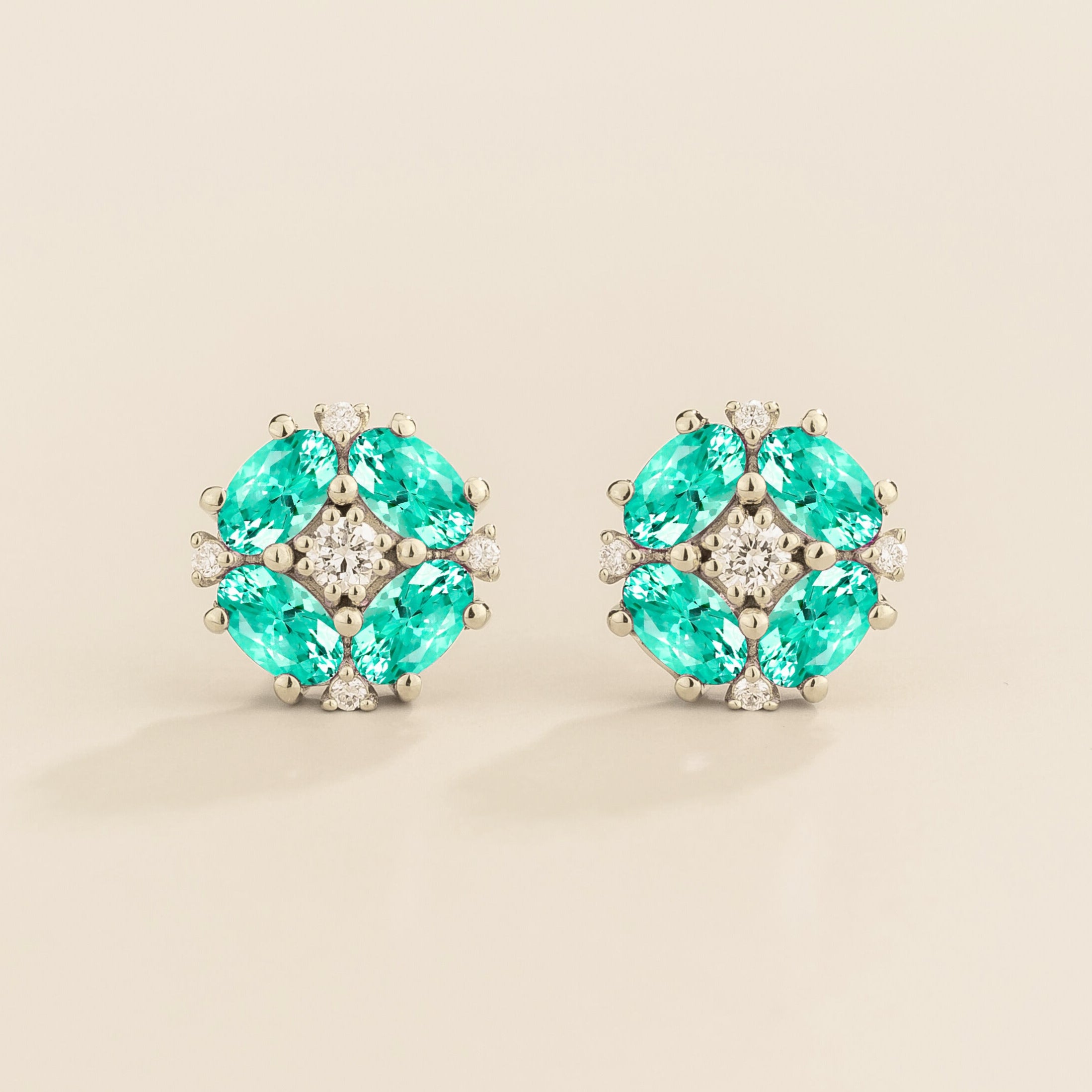 A pair of white gold earrings featuring a central round diamond with oval paraiba sapphire petals arranged around it.