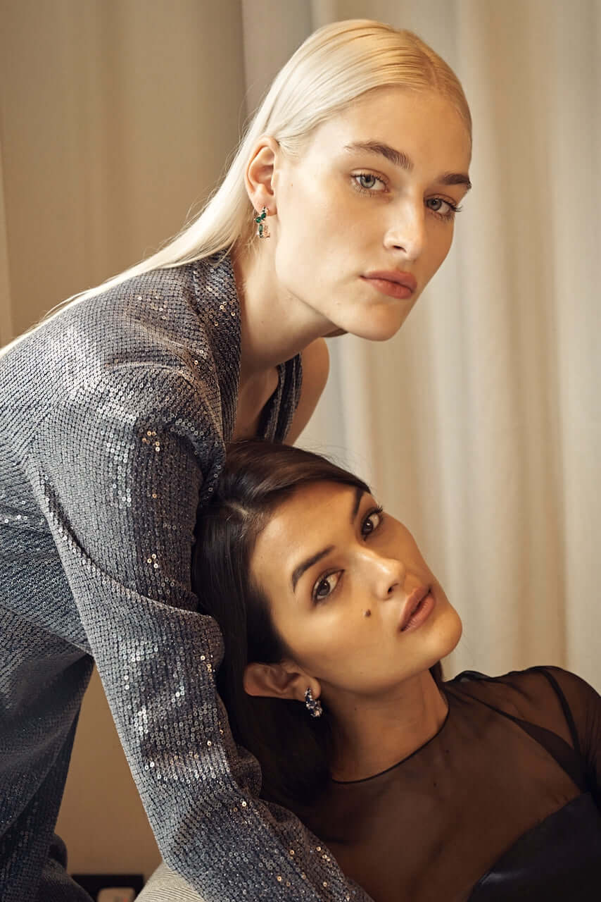 Two models in night cocktail dresses. One is wearing medium hoop Lanna earrings with lab grown emerald and diamond. Another wearing medium hoop Lanna earrings with lab grown blue sapphire and diamond.