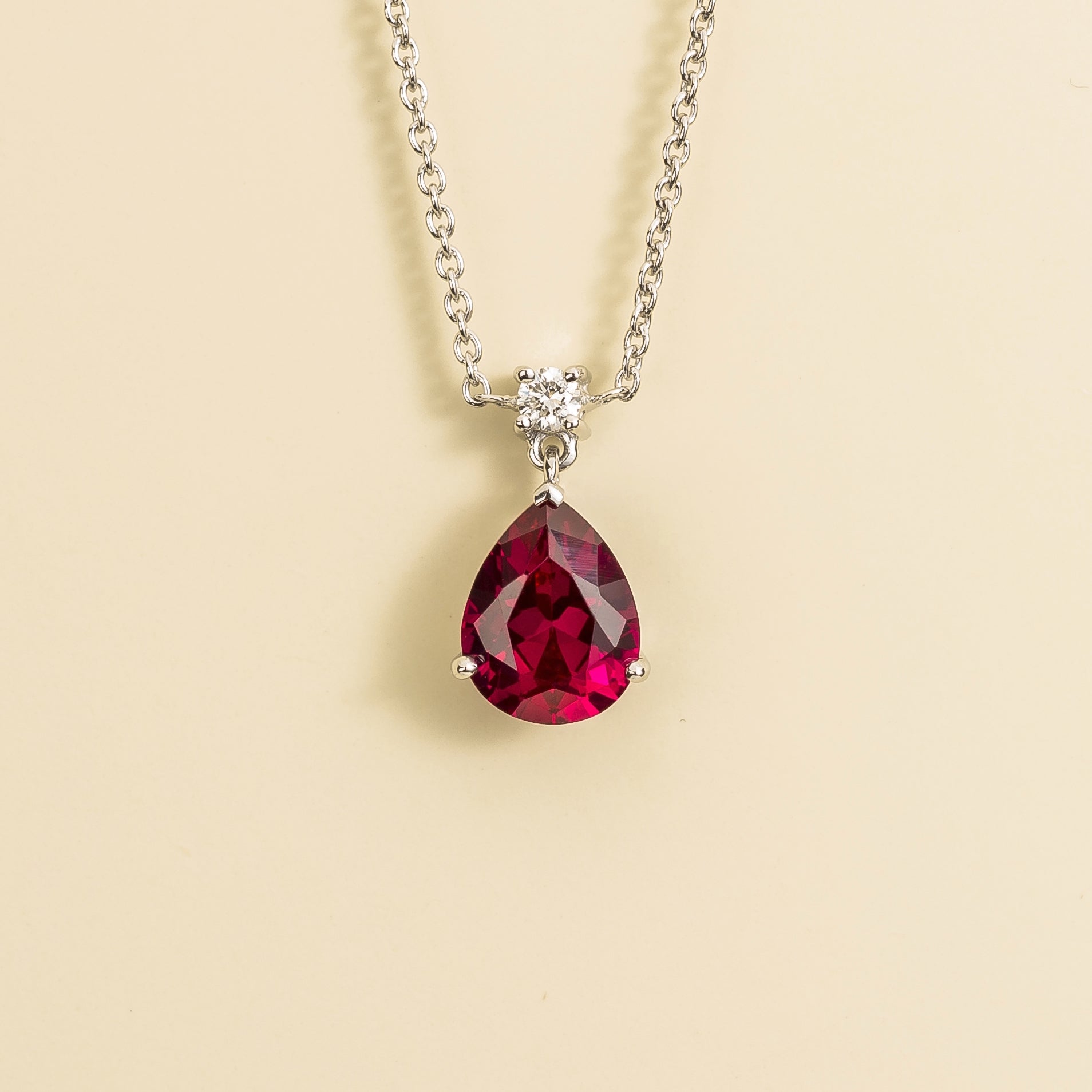 A medium-sized classic pendant necklace in white gold, featuring a pear shape ruby centerpiece and smaller round diamonds, displayed against a neutral background.