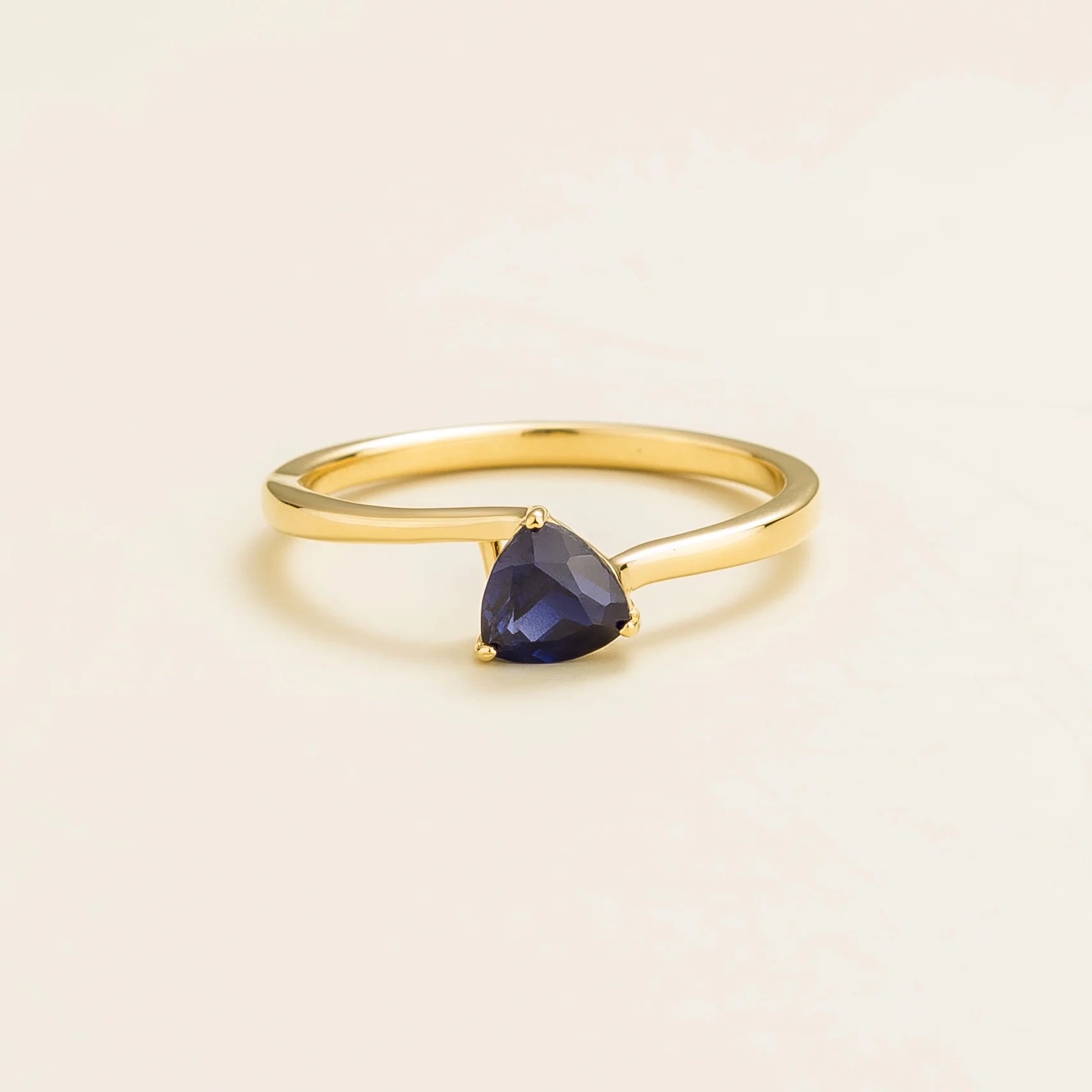A solitaire gold ring with a trillion-cut blue sapphire in a curved modern setting on a plain background.
