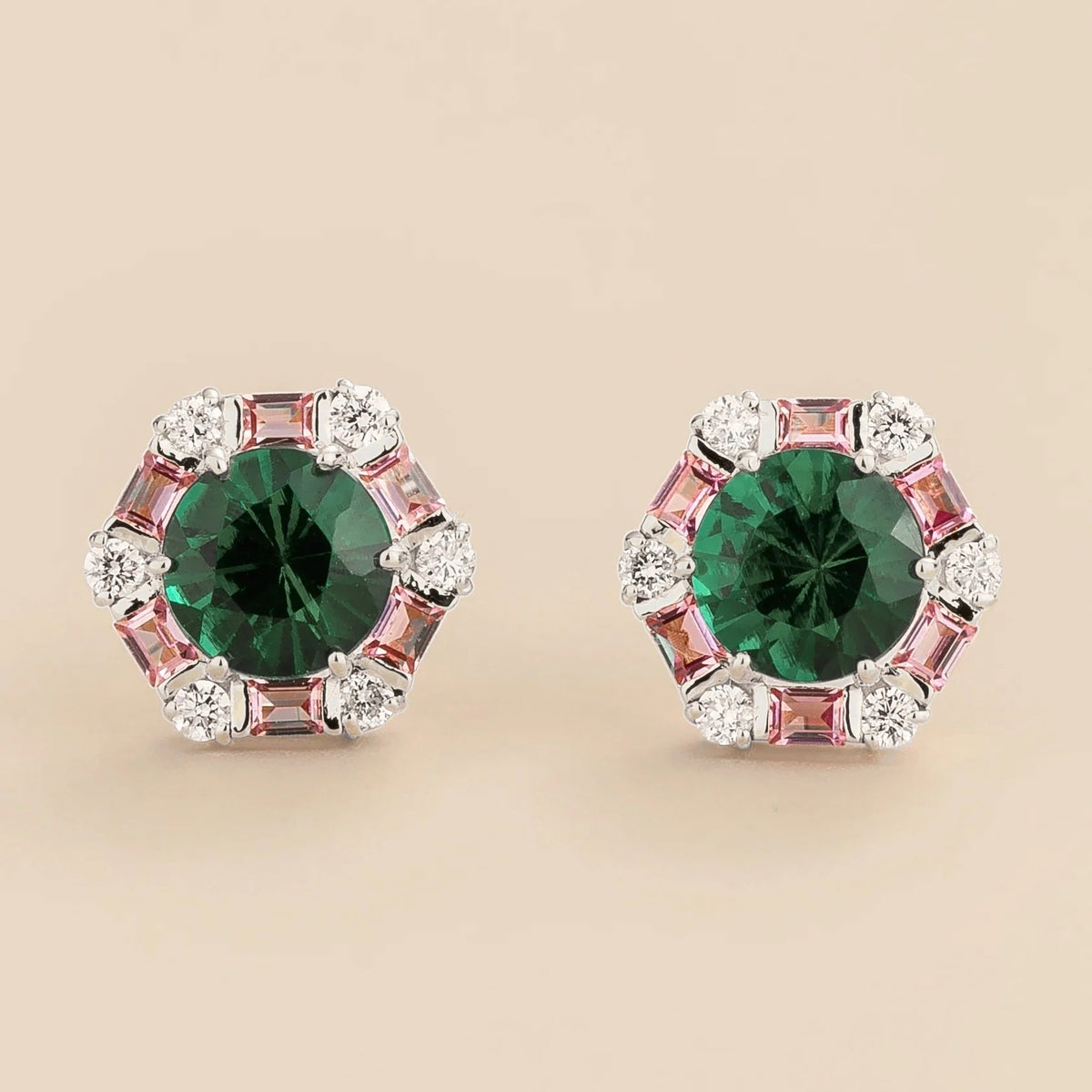White gold stud earrings featuring round emeralds surrounded by pink sapphire baguettes and diamond halo accents.