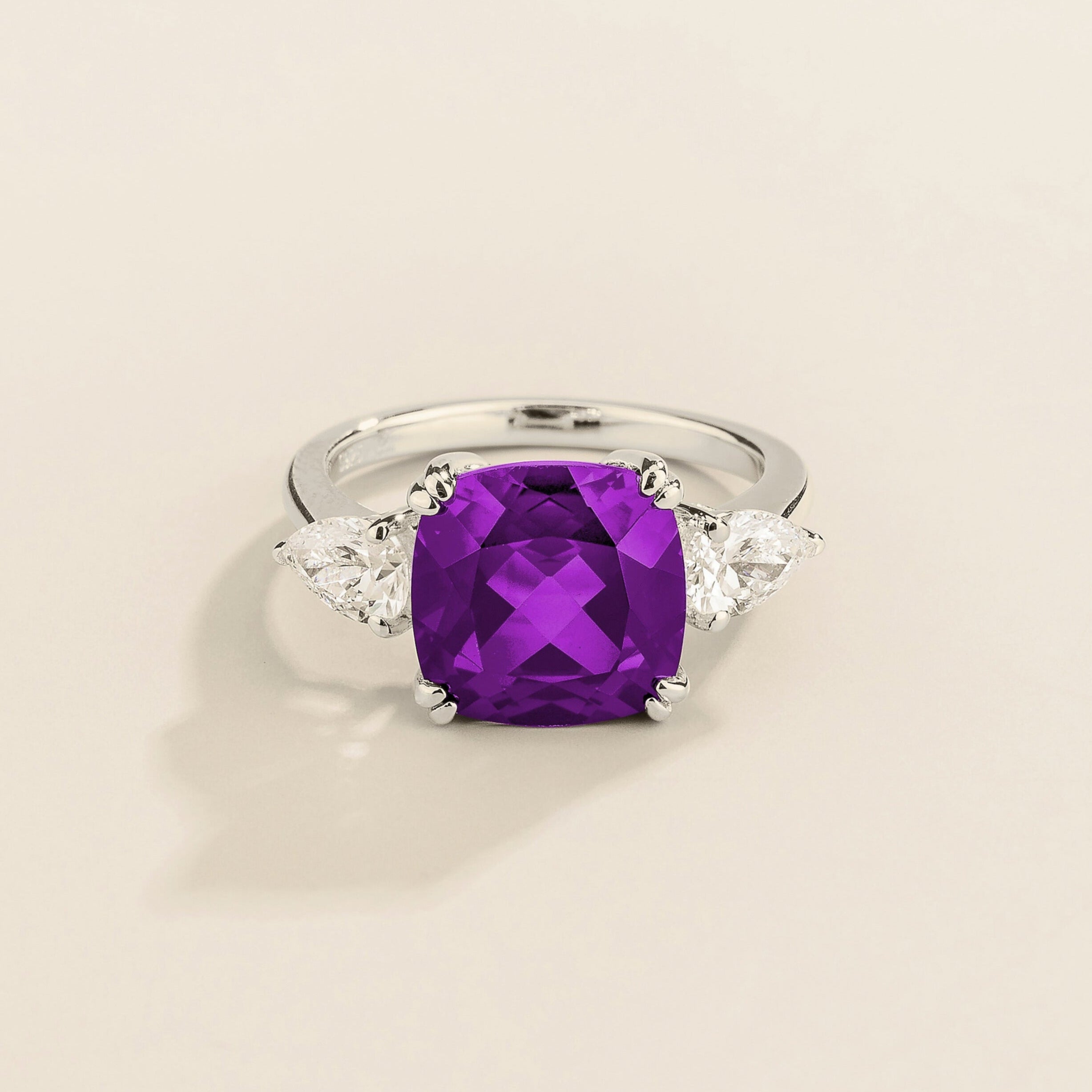 White gold ring with cushion cut purple sapphire gemstone and pear side diamonds on a beige background