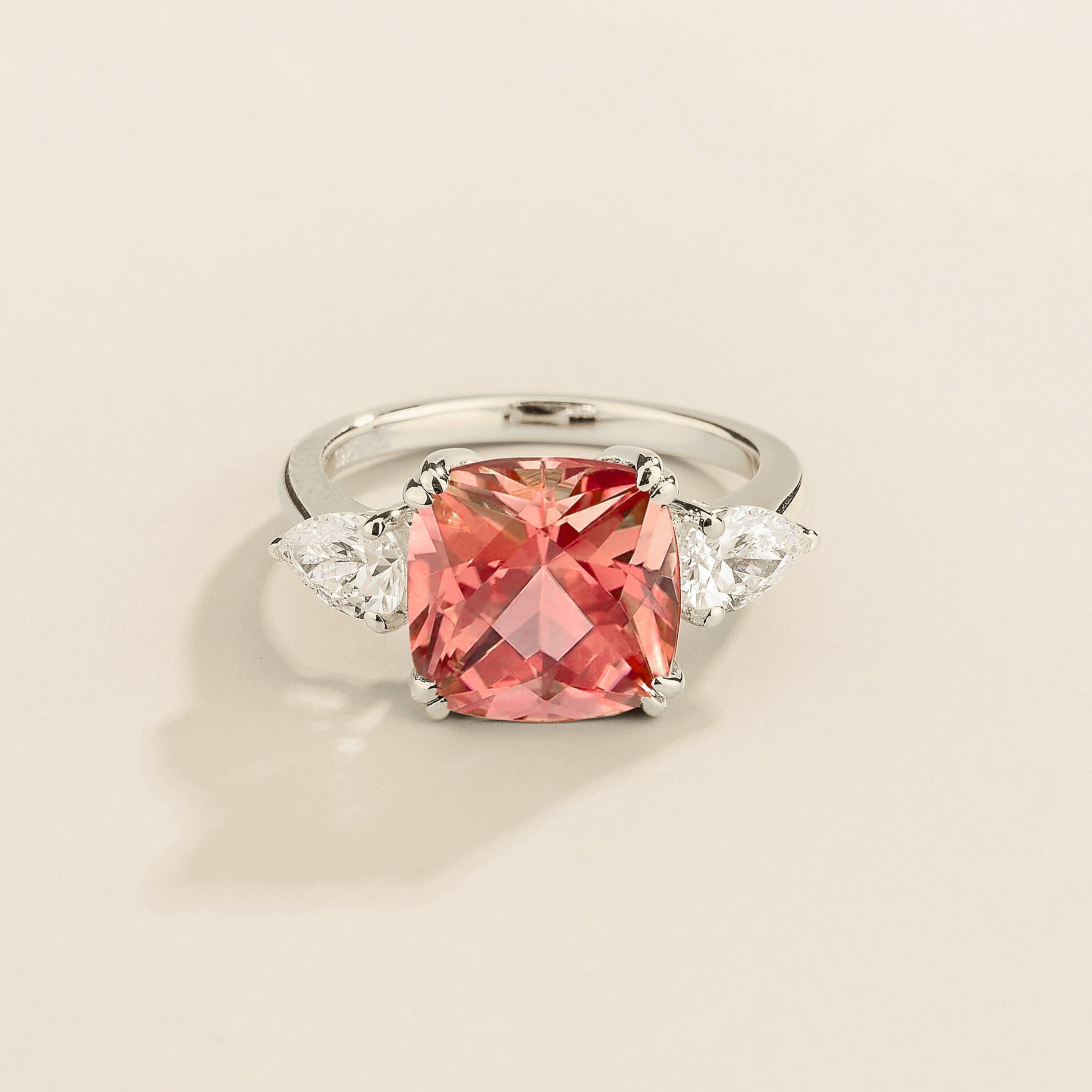 White gold ring with cushion cut padparadscha gemstone and pear side diamonds on a beige background