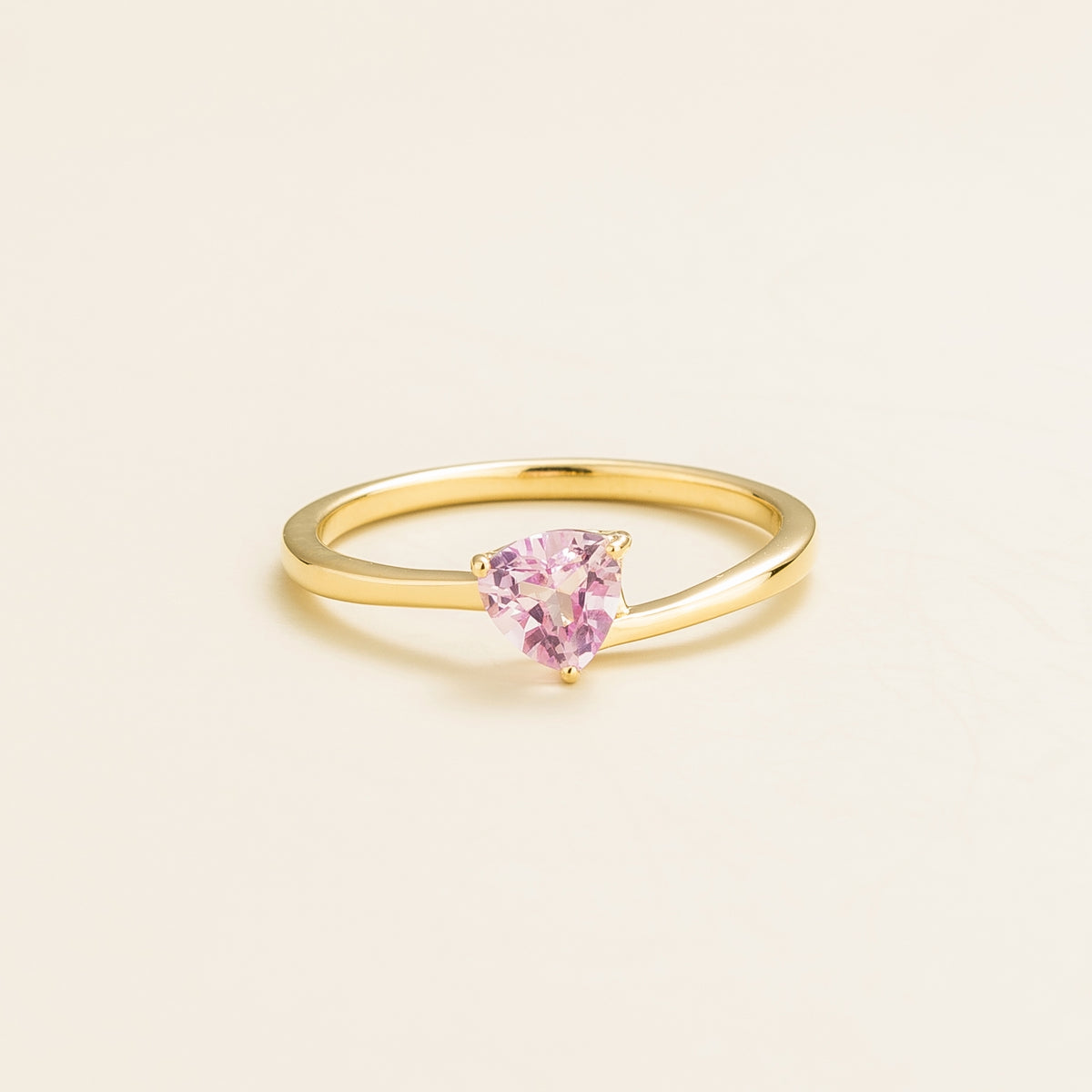 A solitaire gold ring with a trillion-cut pink sapphire in a curved modern setting on a plain background.