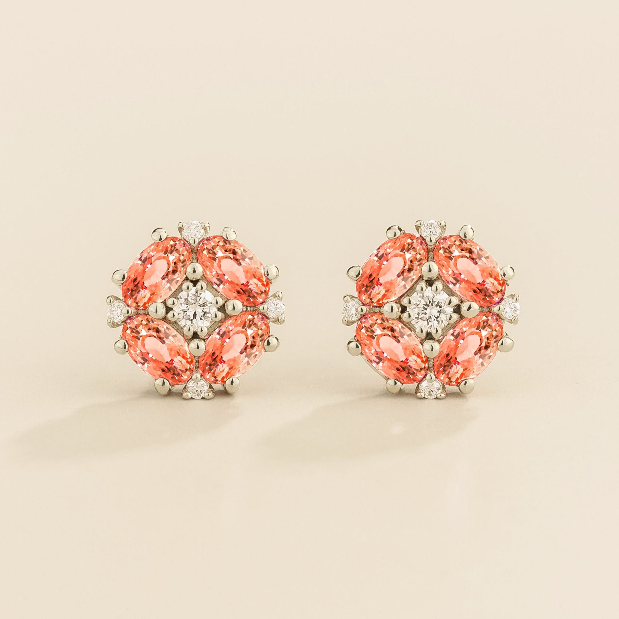 A pair of white gold earrings featuring a central round diamond with oval padparadscha sapphire petals arranged around it.