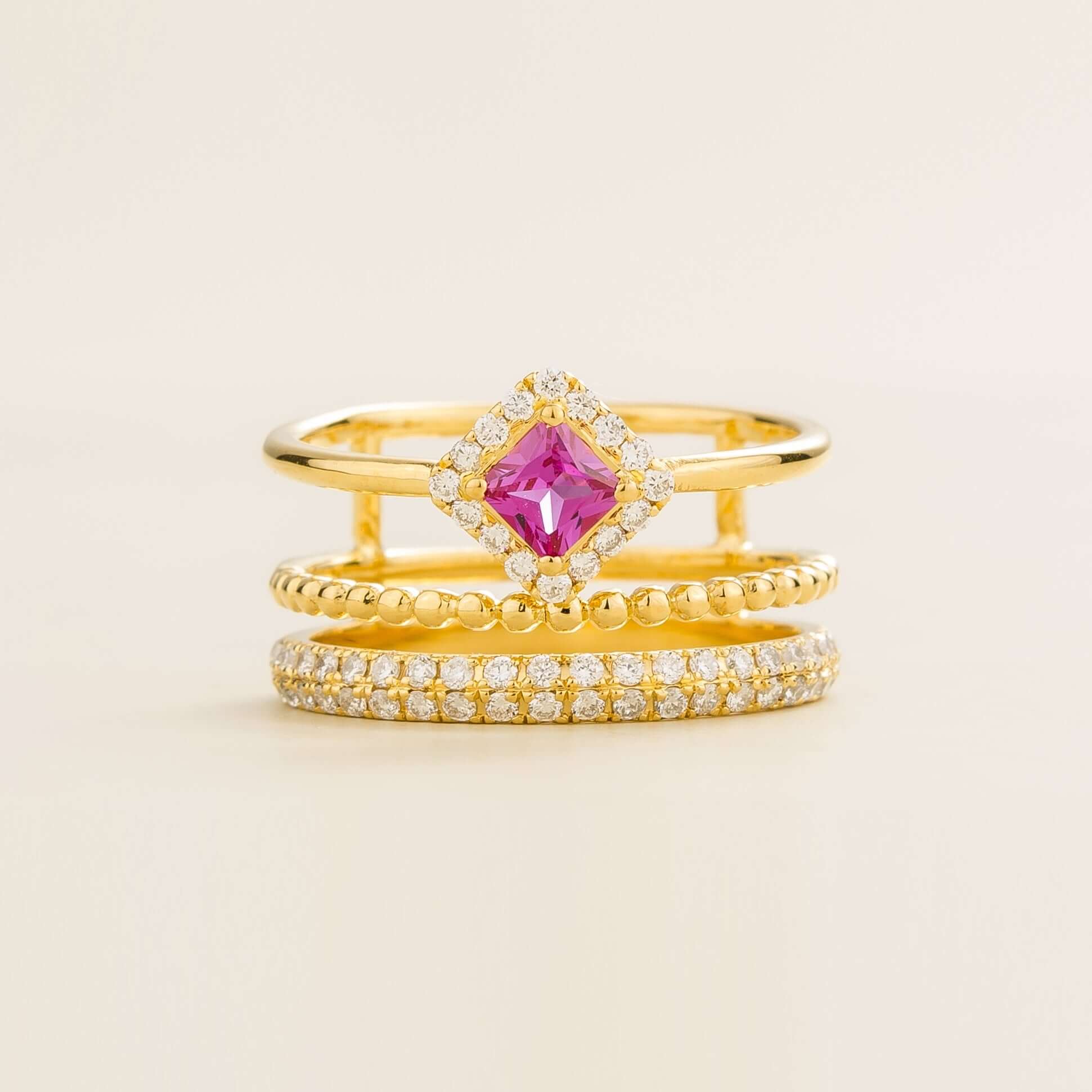 A stack of three gold rings, with the top ring featuring a central princess cut pink sapphire surrounded by small diamonds, and the bottom two bands encrusted with smaller diamonds pavé.