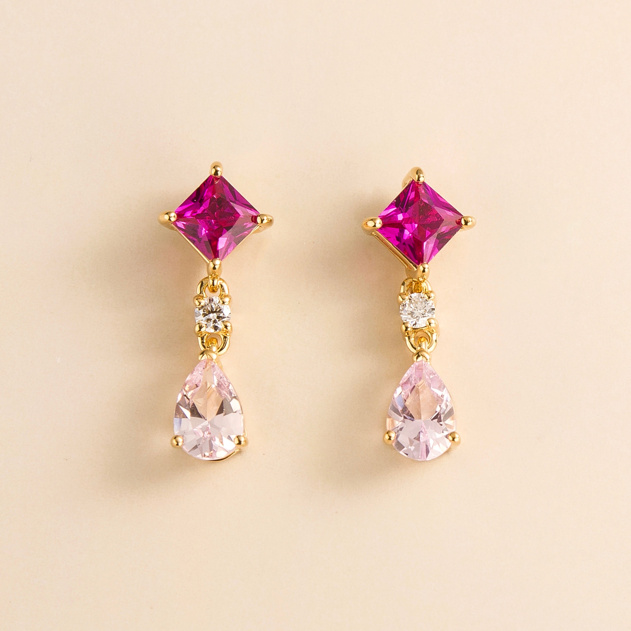A pair of gold drop earrings with one side featuring a square pink sapphire and the other side a pear-shaped pink sapphire, both with smaller round gold accents.