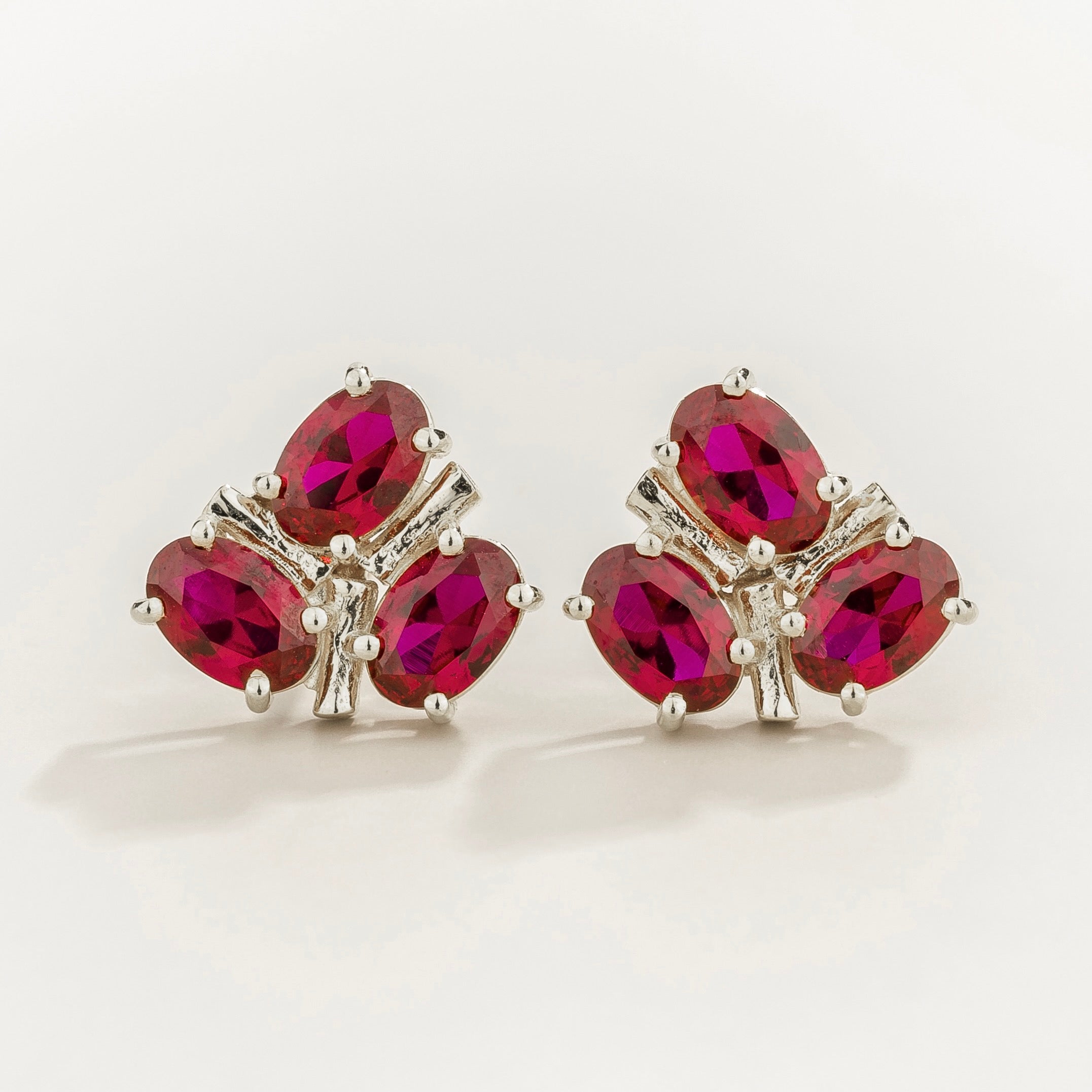 White gold earrings with 3 oval ruby cluster gemstones on a beige background