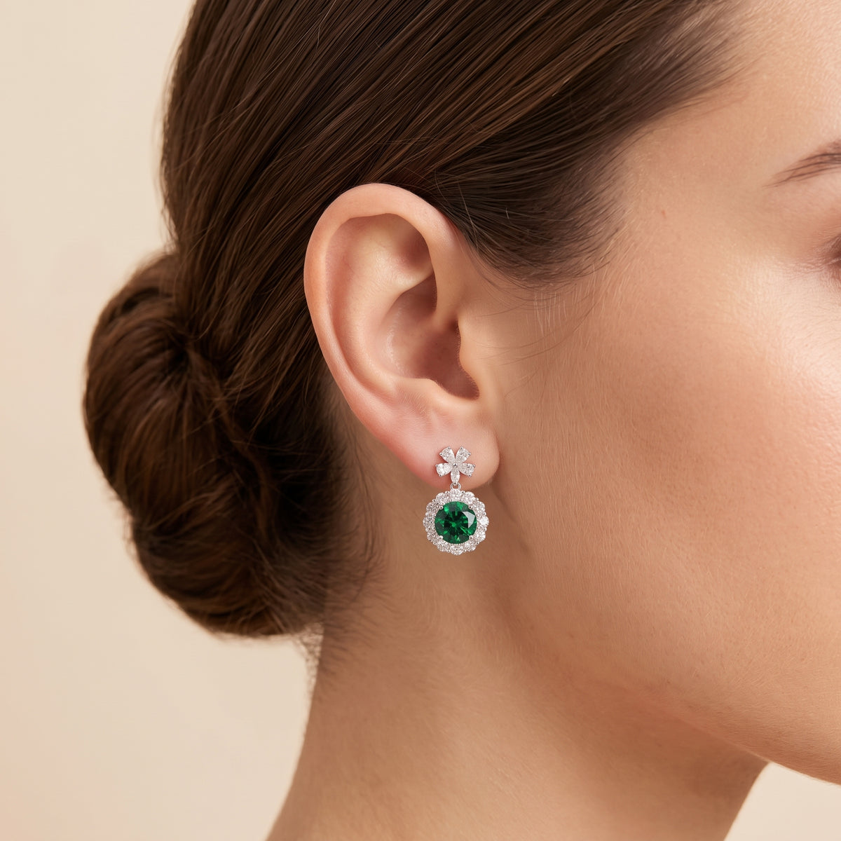 A model is wearing The Antoinette Round Emerald & Diamond Halo Pendant Earrings with Flower Bail