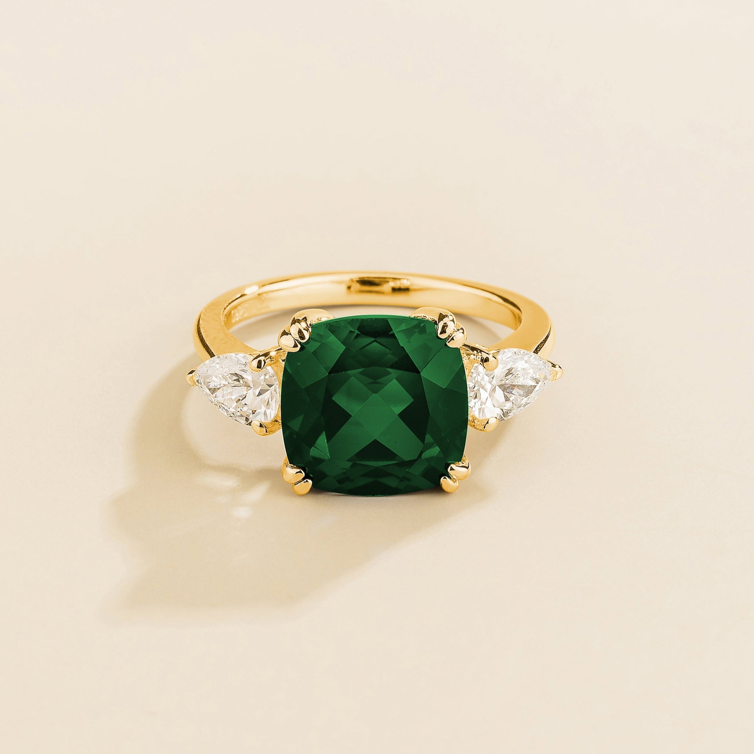 Gold ring with cushion cut emerald gemstone and pear side diamonds on a beige background