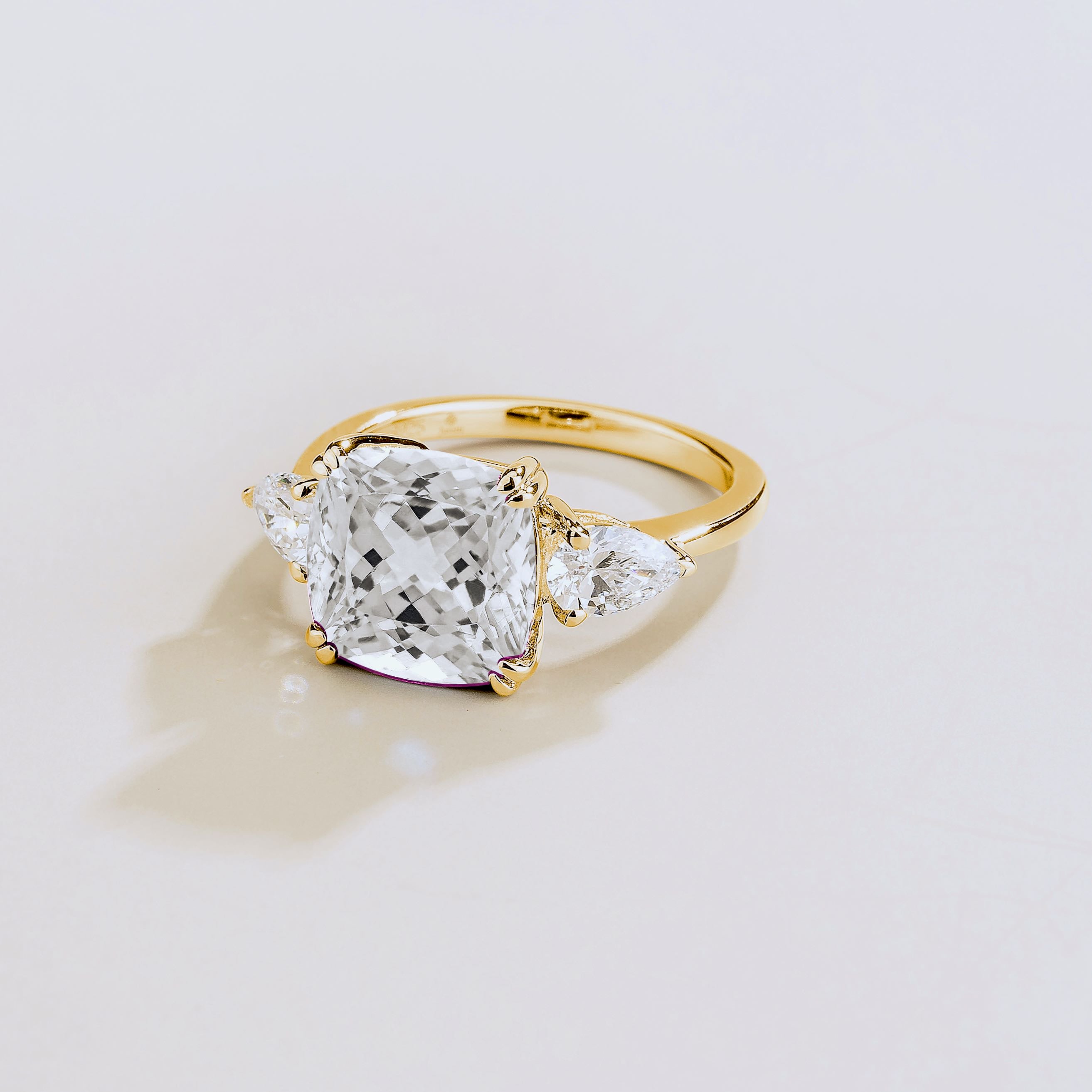 Gold ring with large cushion cut diamond gemstone and pear side diamonds on a beige background
