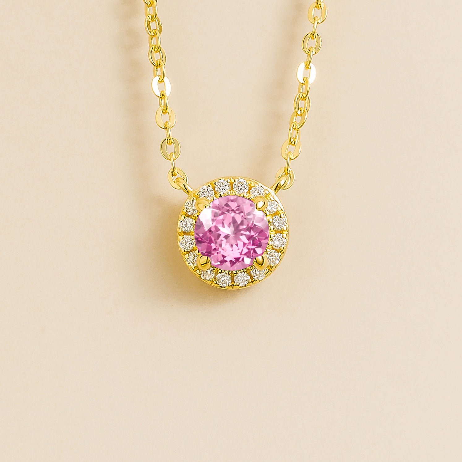 A gold gold necklace with a halo design featuring a round pink sapphire centerpiece surrounded by smaller diamonds, displayed against a light background.