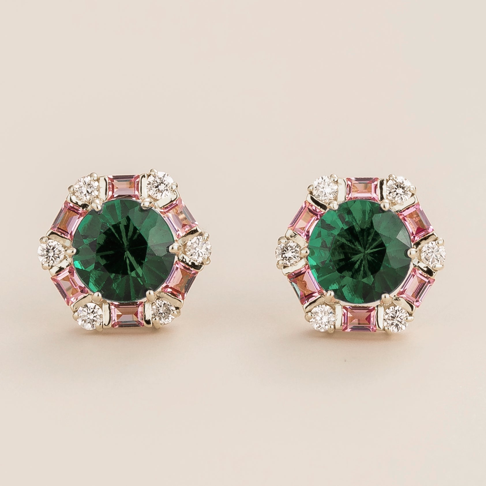White gold stud earrings featuring round emeralds surrounded by pink sapphire baguettes and diamond halo accents.