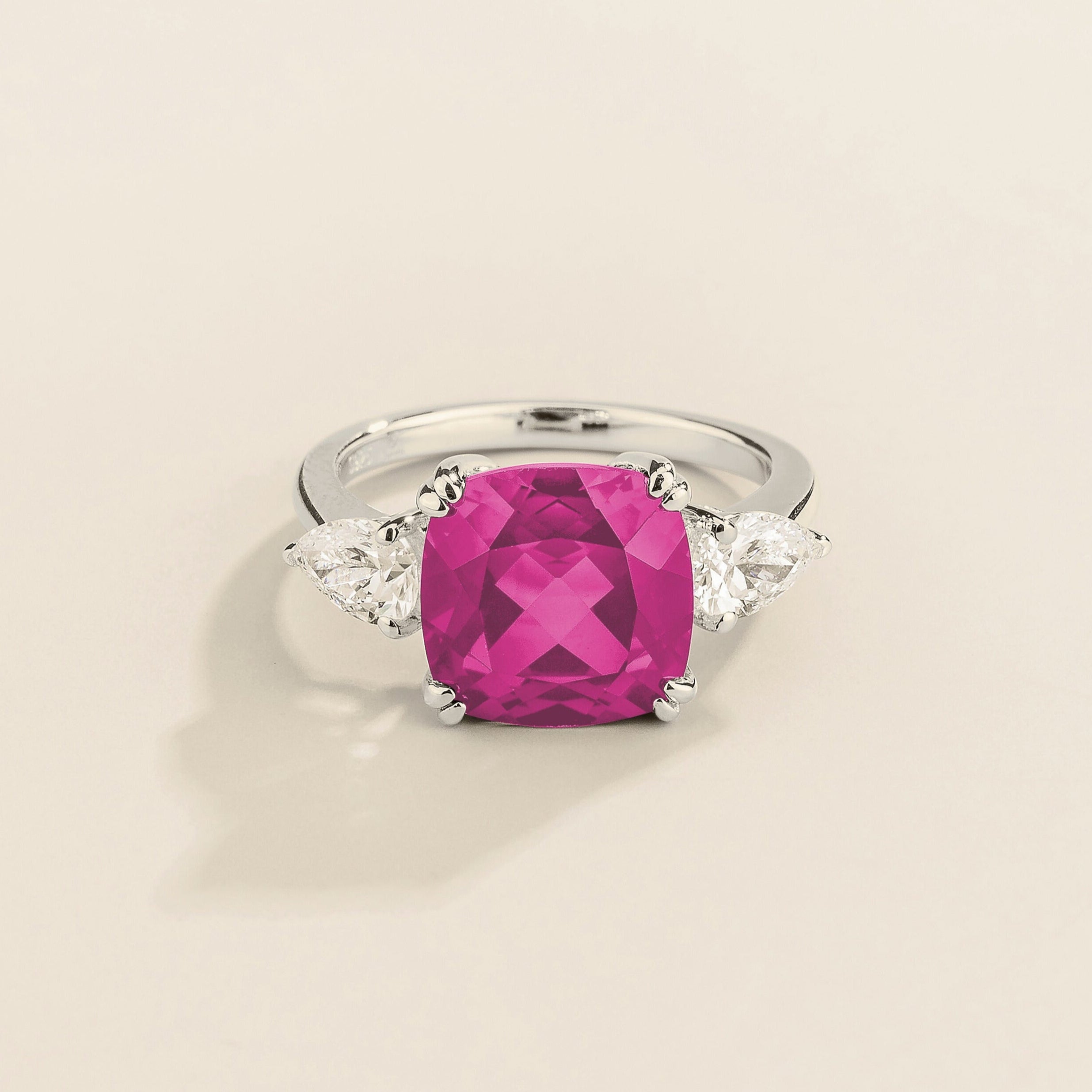 White gold ring with cushion cut vivid pink sapphire gemstone and pear side diamonds on a beige background