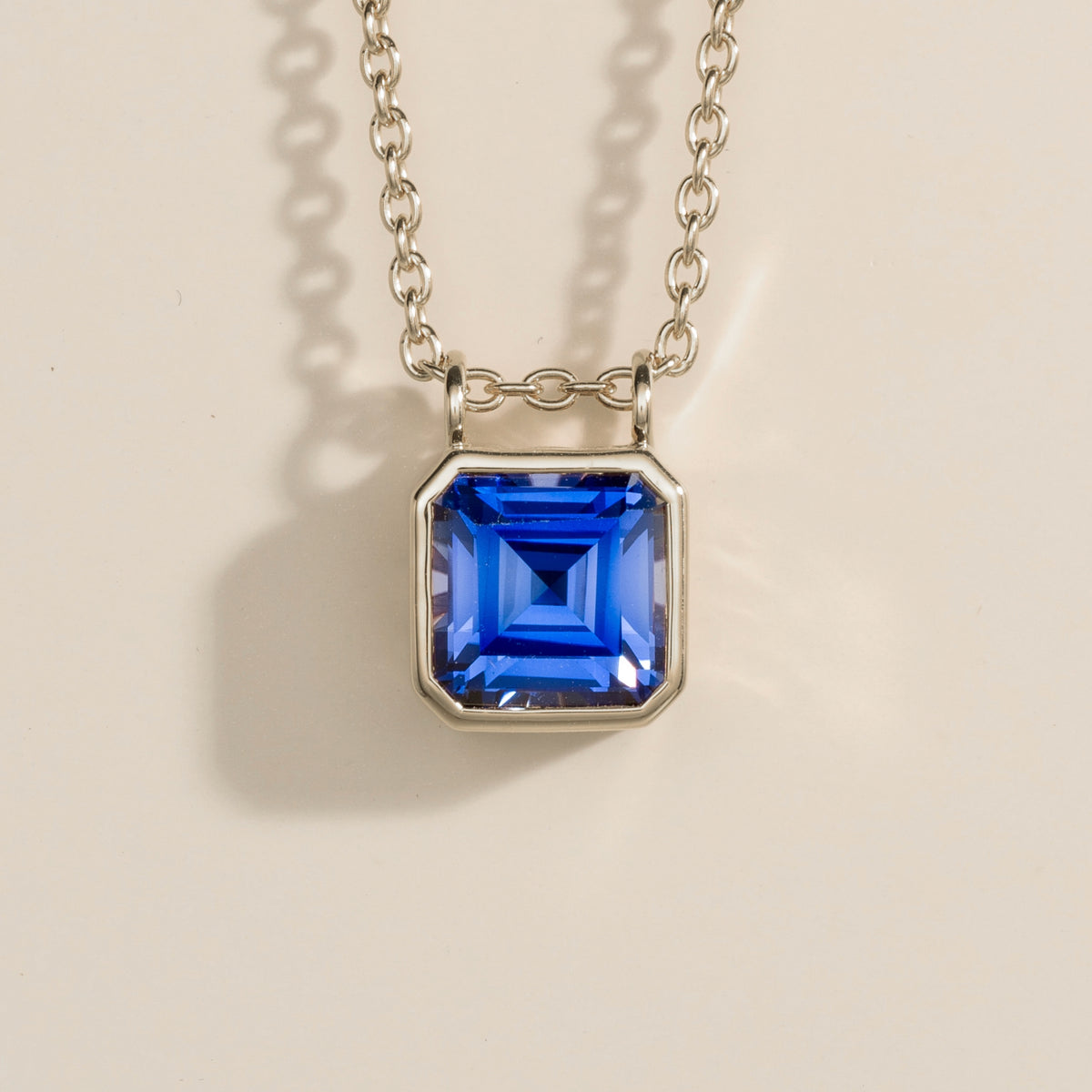 A white gold pendant necklace with a bezel set blue sapphire asscher cut on a light background.