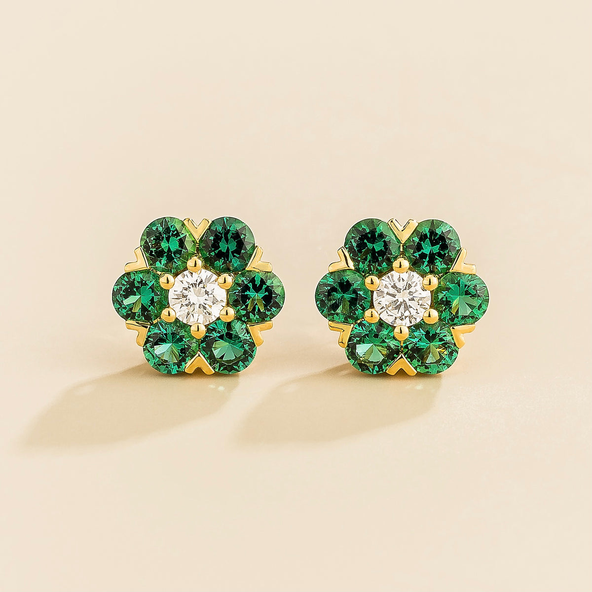 A pair of gold earrings featuring a central diamond with emerald accents arranged in a floral daisy pattern.