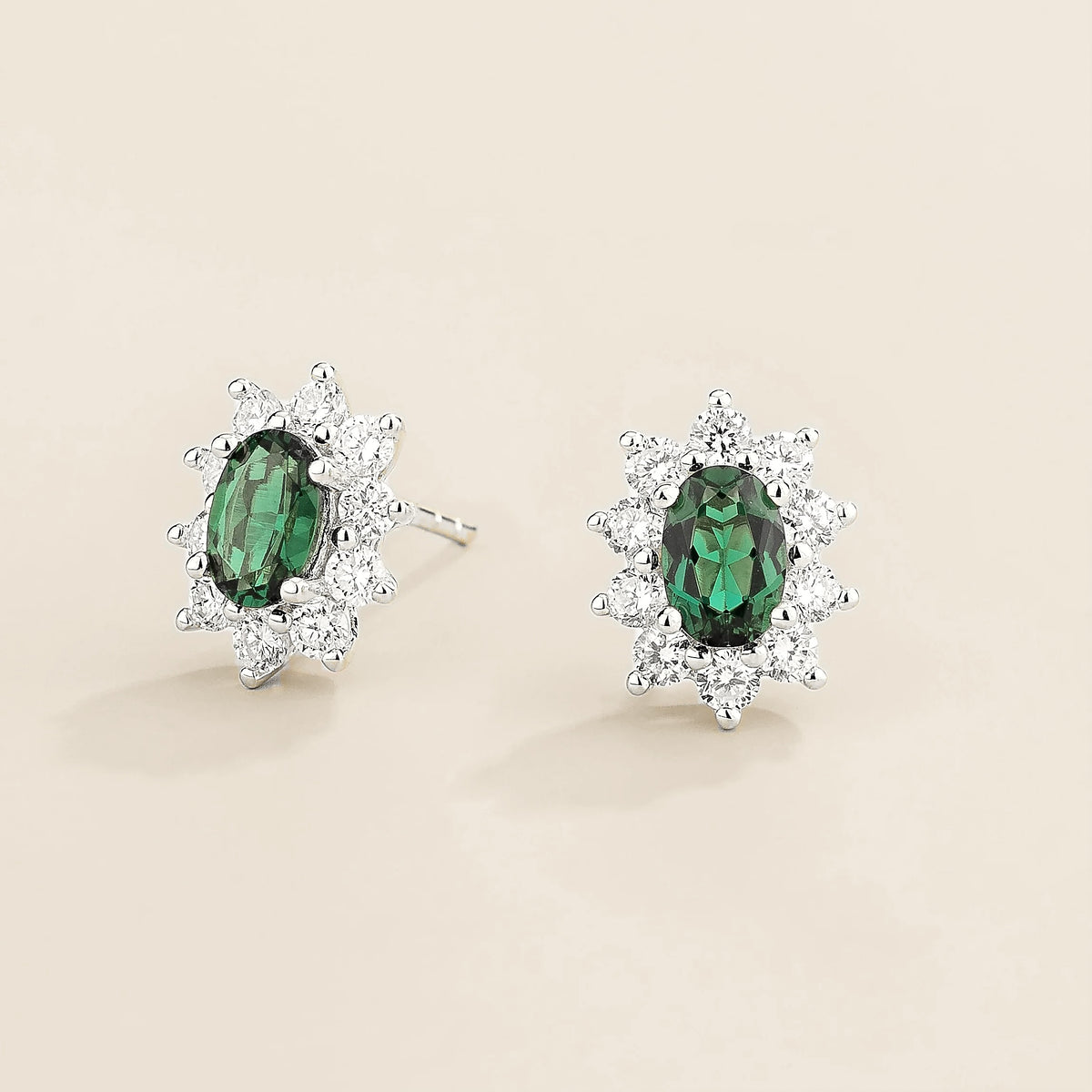 Pair of earrings with oval green emerald gemstones and diamonds halo on a light background