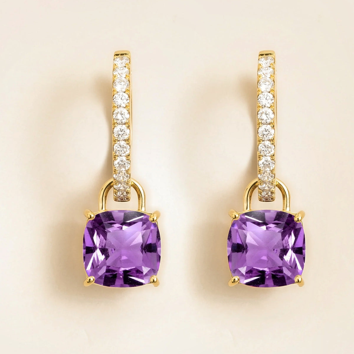 A pair of gold hoop earrings with cushion purple sapphire drops and diamonds huggie hoop, set against a light beige background.