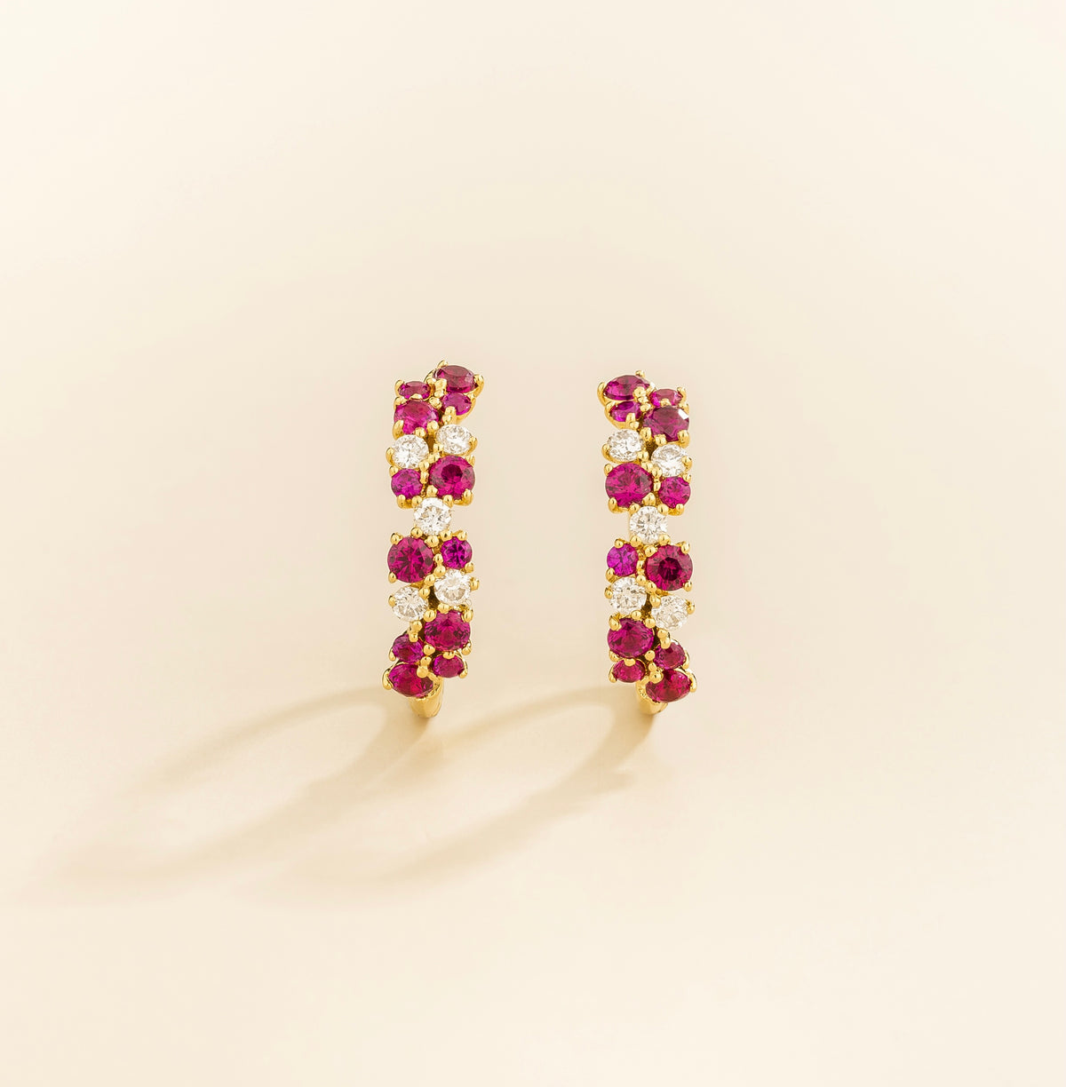Pair of gold huggie hoop earrings with ruby and diamond gemstones on a beige background