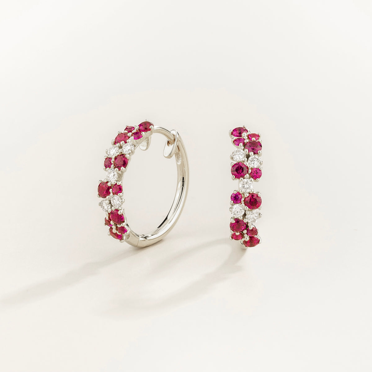 A pair of white gold huggie earrings set with cluster of round ruby and diamond accents, displayed against a neutral background.