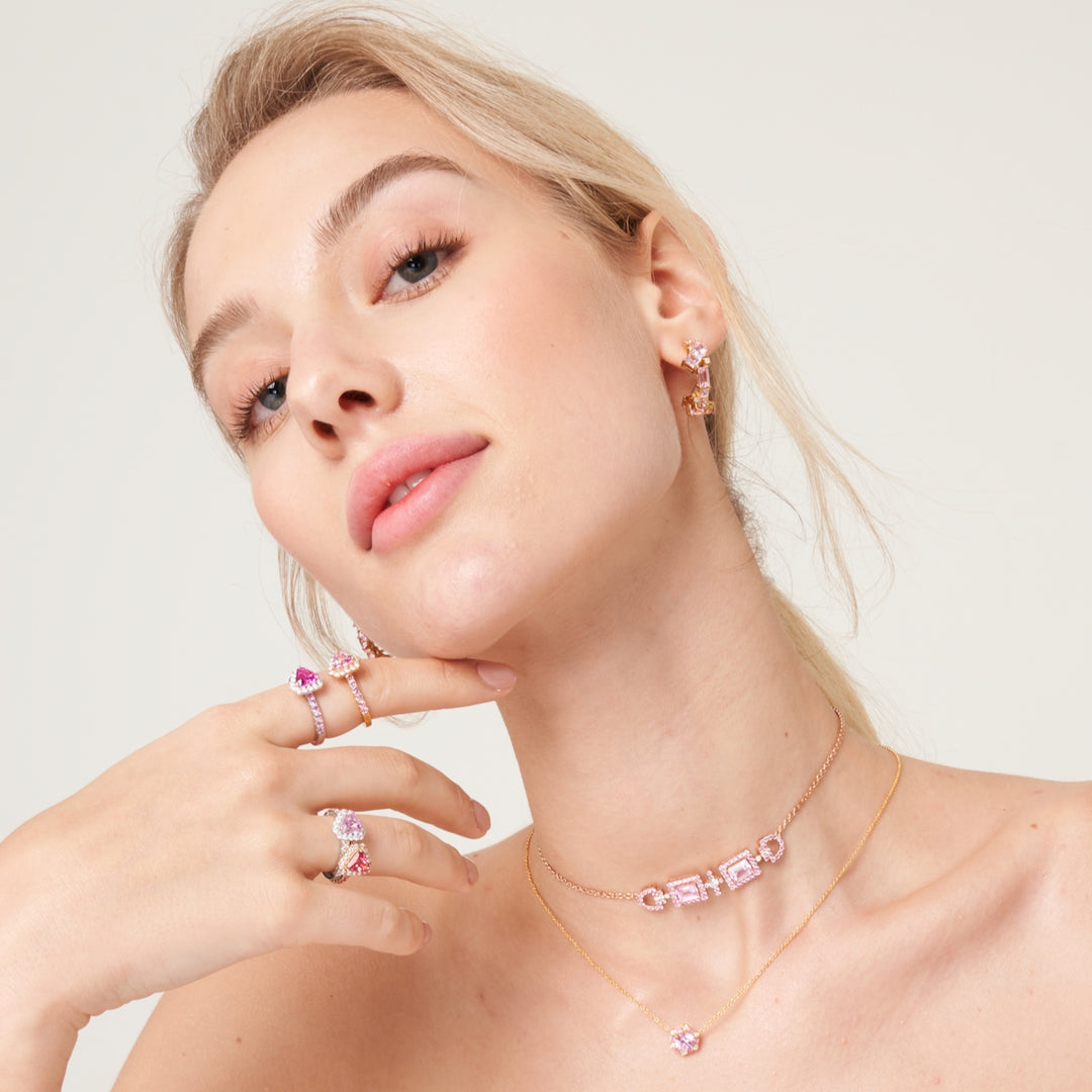 A model wearing medium hoop Lanna earrings, triangle Diana rings, hexagon melba necklace and cross baguette Ciceris necklace. The pieces are set with lab grown diamond and pink sapphire gem stones.